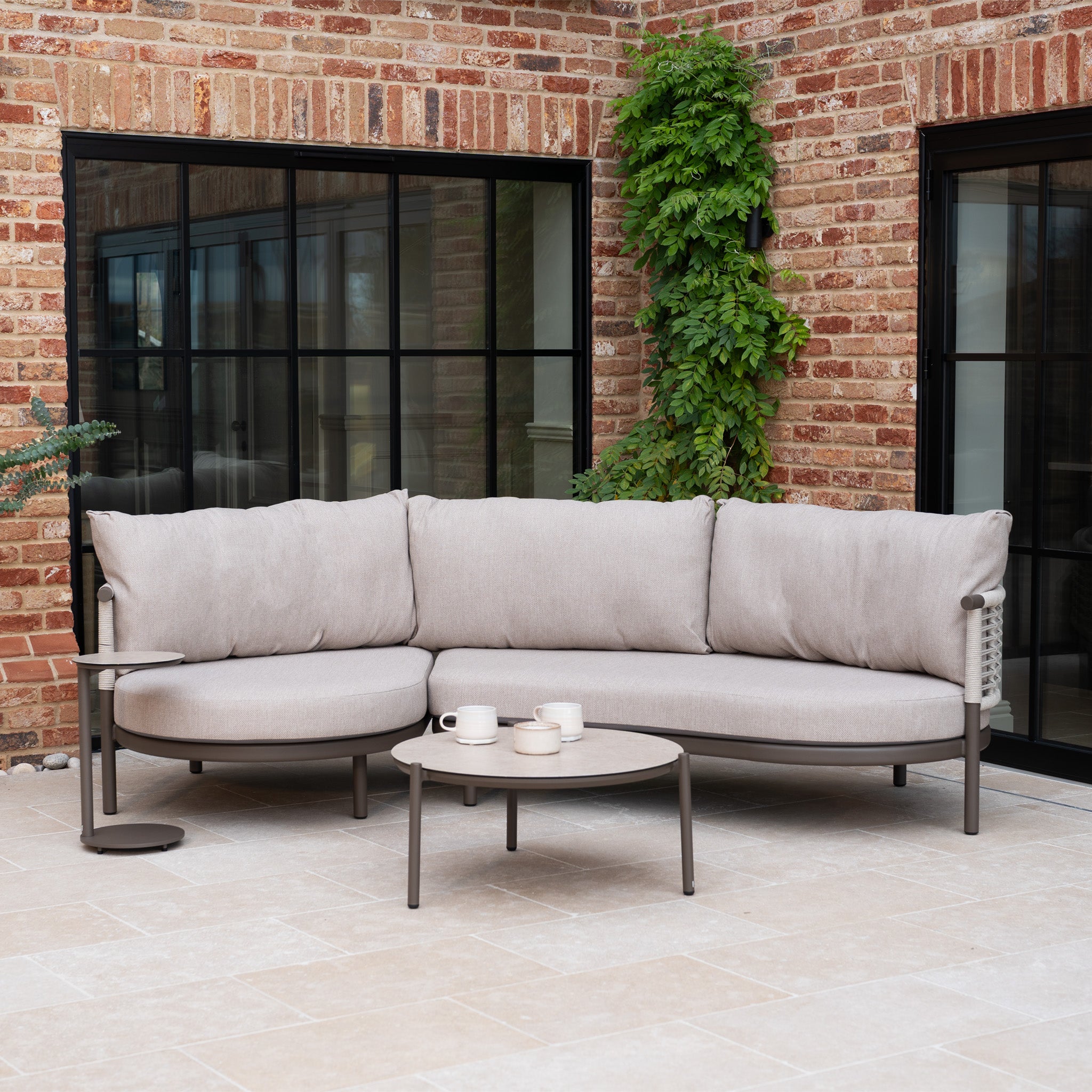 Outdoor patio with a Sutera Curved Chaise Sofa Set in Putty, all-weather cushions, two tables, brick walls, large windows, and greenery.