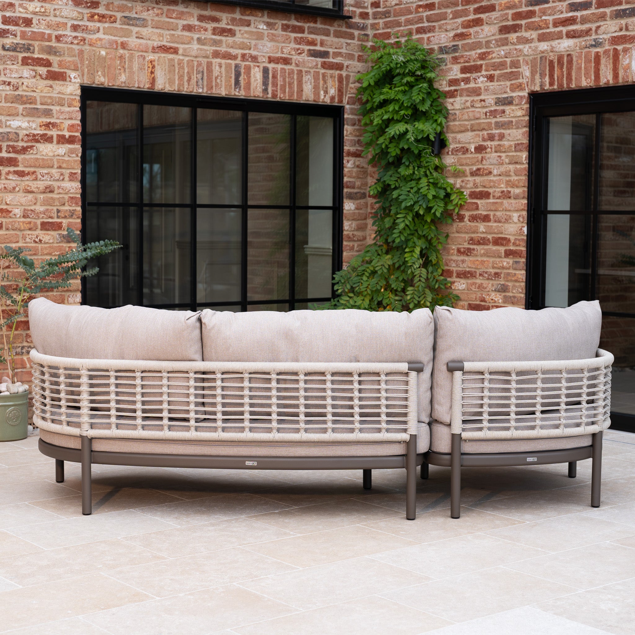 The Sutera Curved Chaise Sofa Set in Putty features woven rope accents and beige all-weather cushions, offering a welcoming spot on the tiled patio beside a brick wall with large windows.