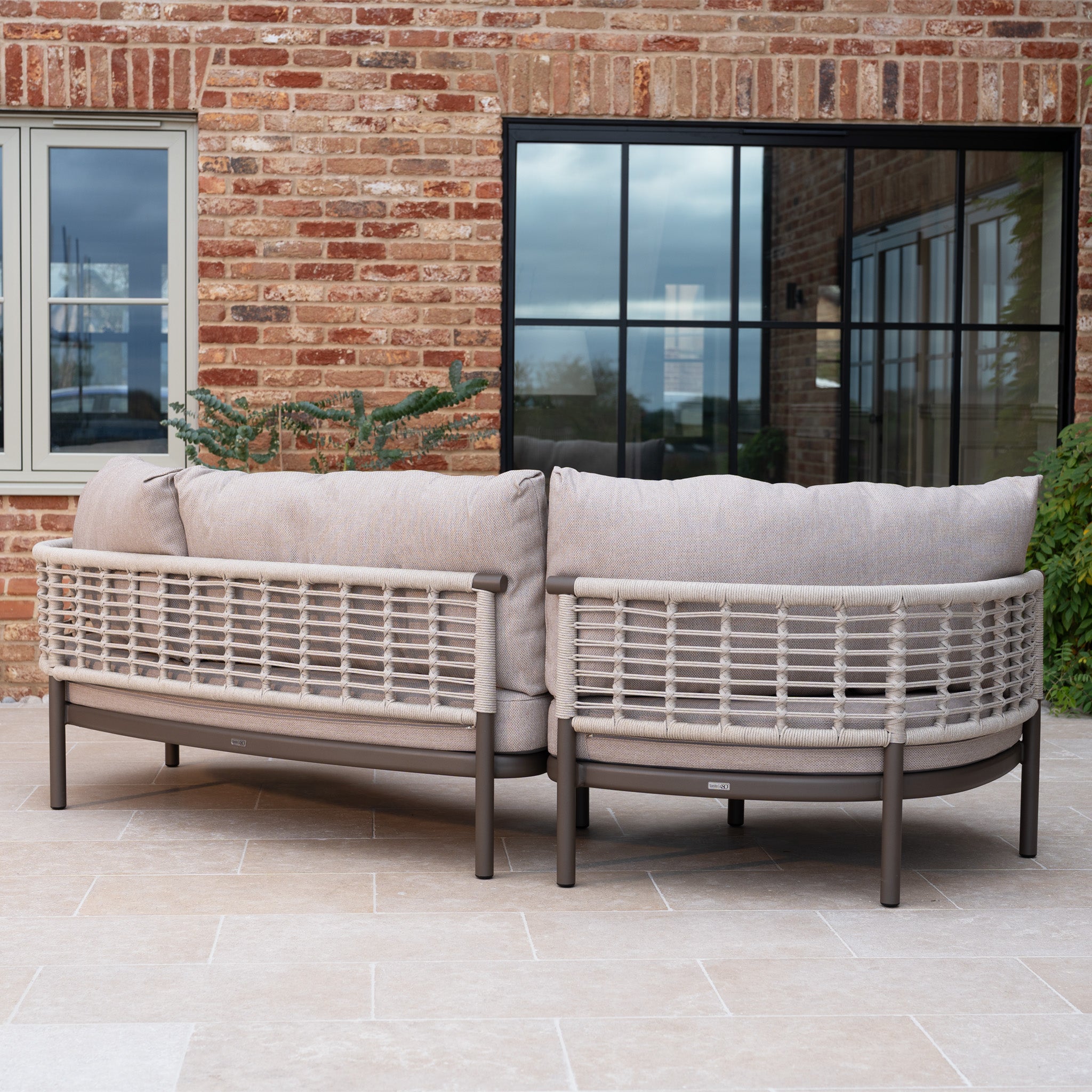 Outdoor patio featuring two curved, cushioned wicker sofas from the Sutera Curved Chaise Sofa Set in Putty, arranged in front of a brick wall and large windows.