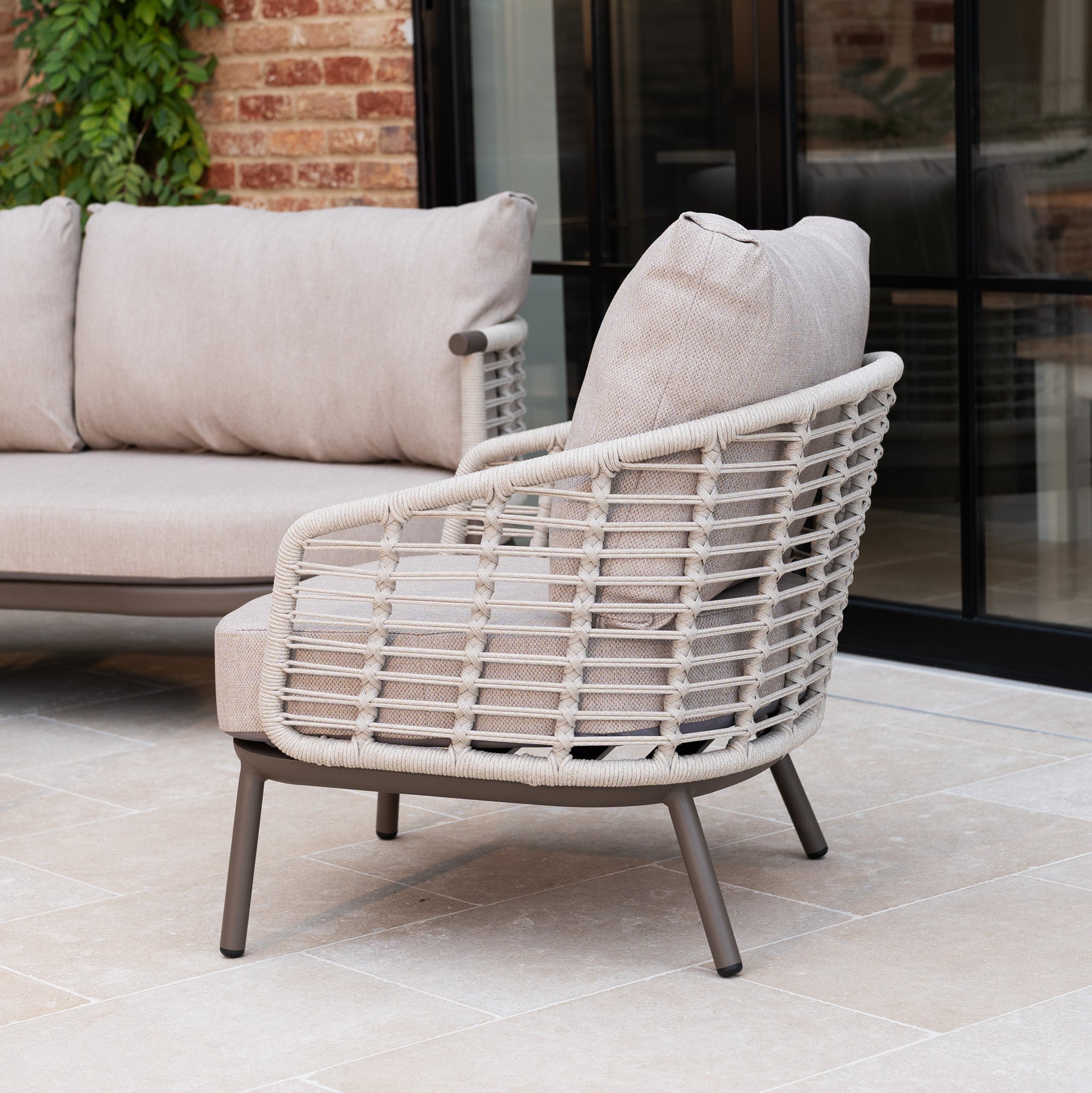Putty outdoor armchair with woven frame and all-weather cushions on a tiled patio, placed next to the Sutera Curved Chaise Sofa Set with Armchair in Putty by glass doors.