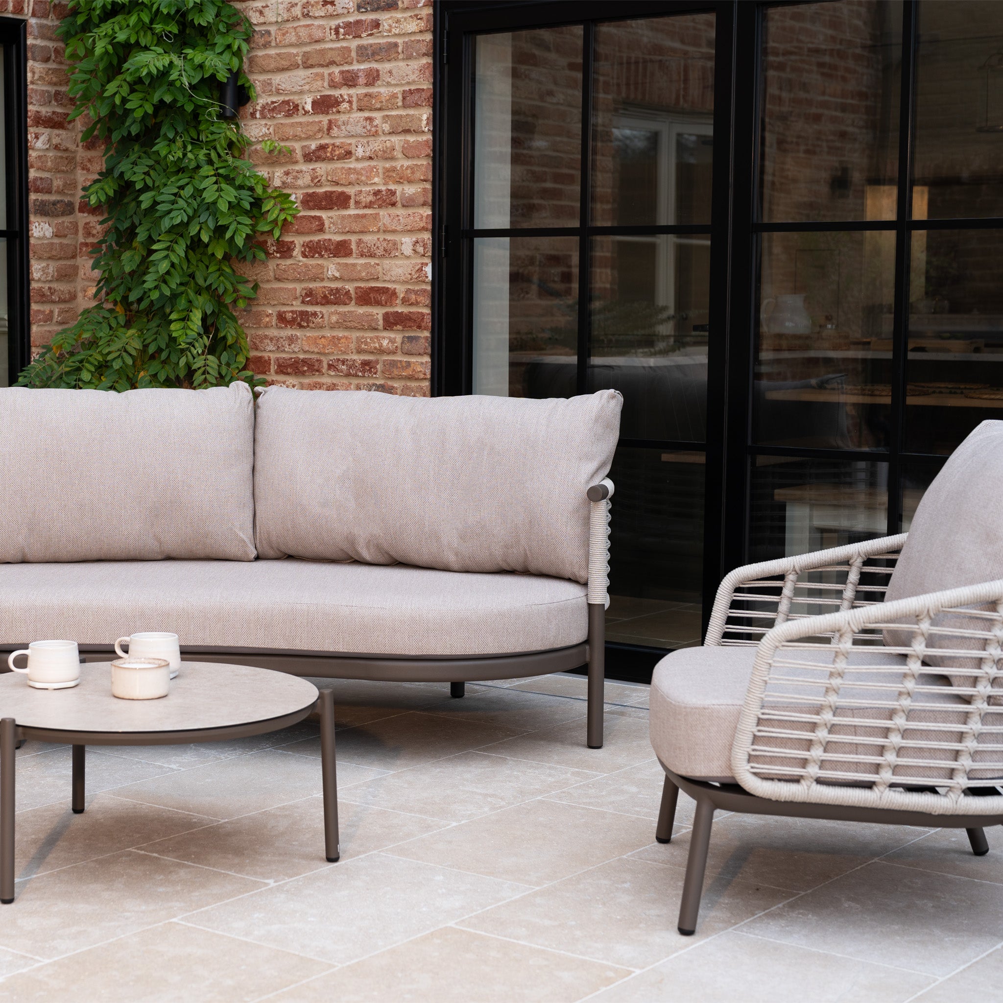 Modern patio with outdoor garden furniture, featuring the Sutera Curved Chaise Sofa Set with Armchair in Putty and a round table on tiled floor by a brick wall draped in green vine.