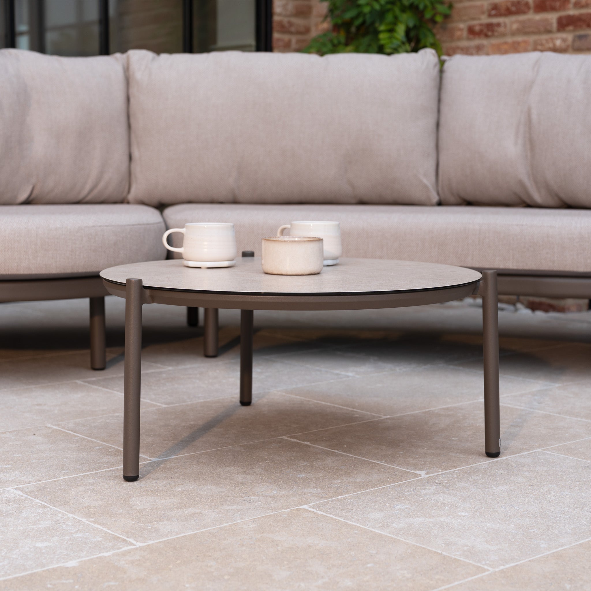 Round coffee table with three mugs in front of the Sutera Curved Chaise Sofa Set with Armchair in Putty, featuring all-weather cushions on a tiled patio—perfect outdoor garden furniture for relaxing.