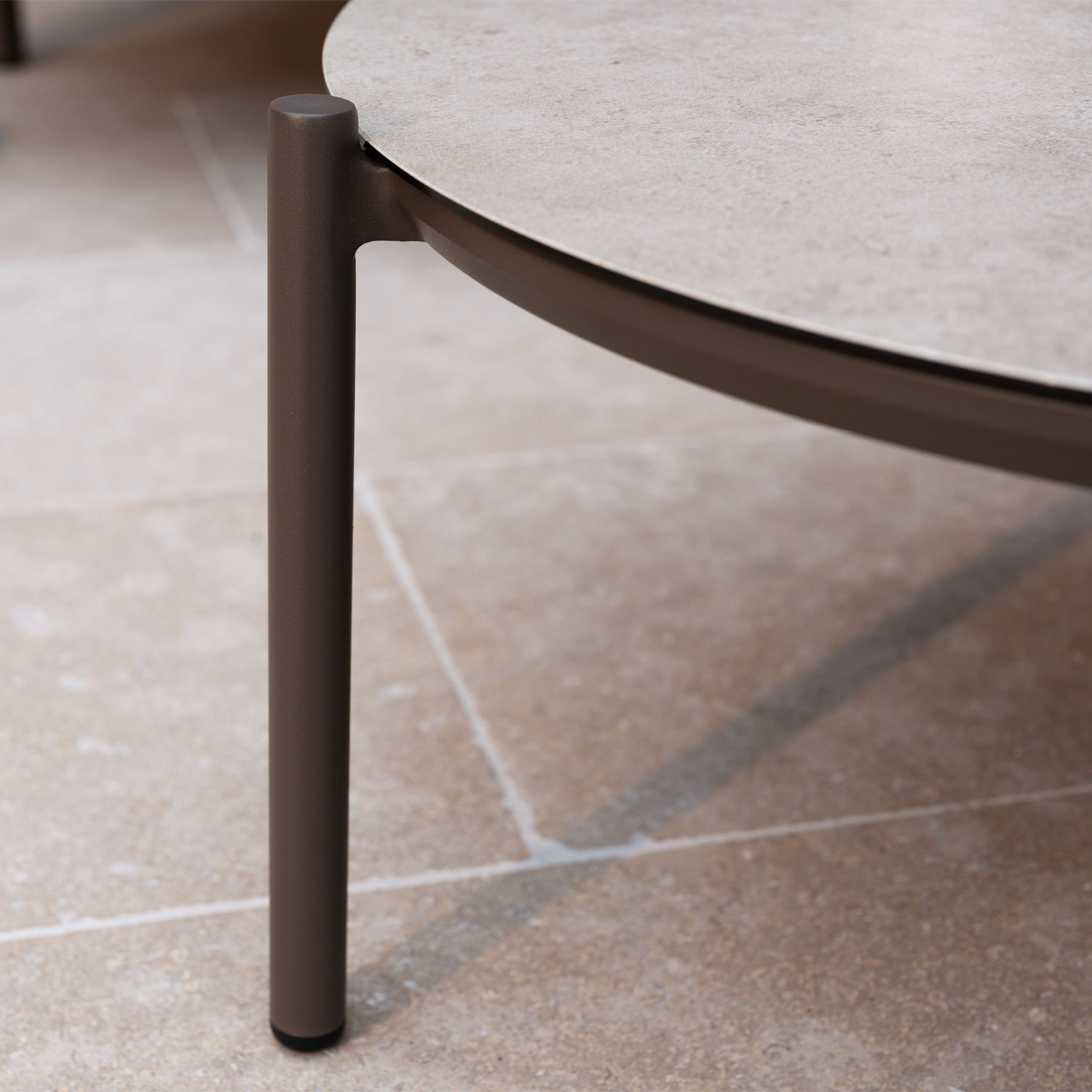 Close-up of a round table with a brown metal leg on a tiled floor, perfect for pairing with outdoor garden furniture like the Sutera Curved Chaise Sofa Set with Armchair in Putty.