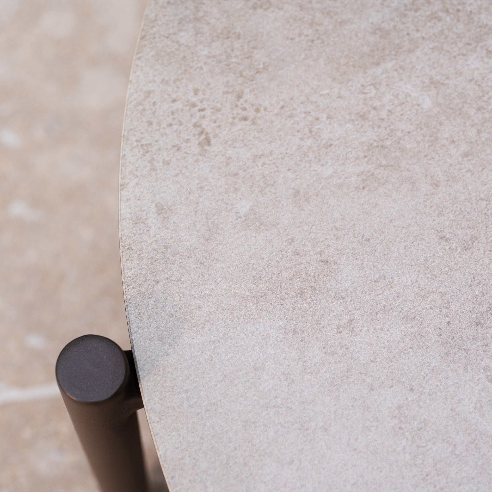Close-up of the Sutera Curved Chaise Sofa Set with Armchair in Putty, featuring a round, light beige stone tabletop and a black metal leg that complements modern outdoor garden furniture.