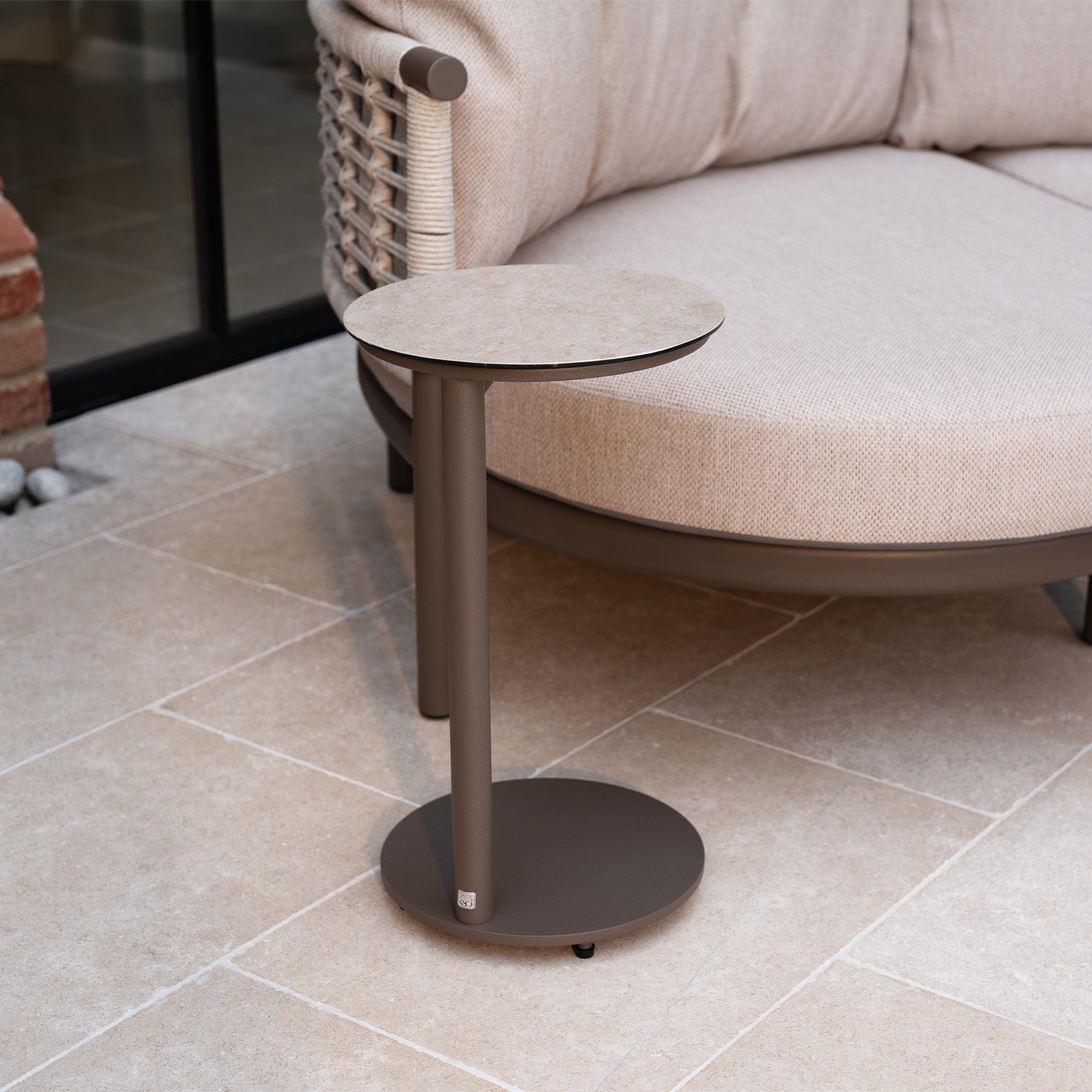 Small round side table with a beige top next to the Sutera Curved Chaise Sofa Set with Armchair in Putty, featuring all-weather cushions, on a tiled patio.