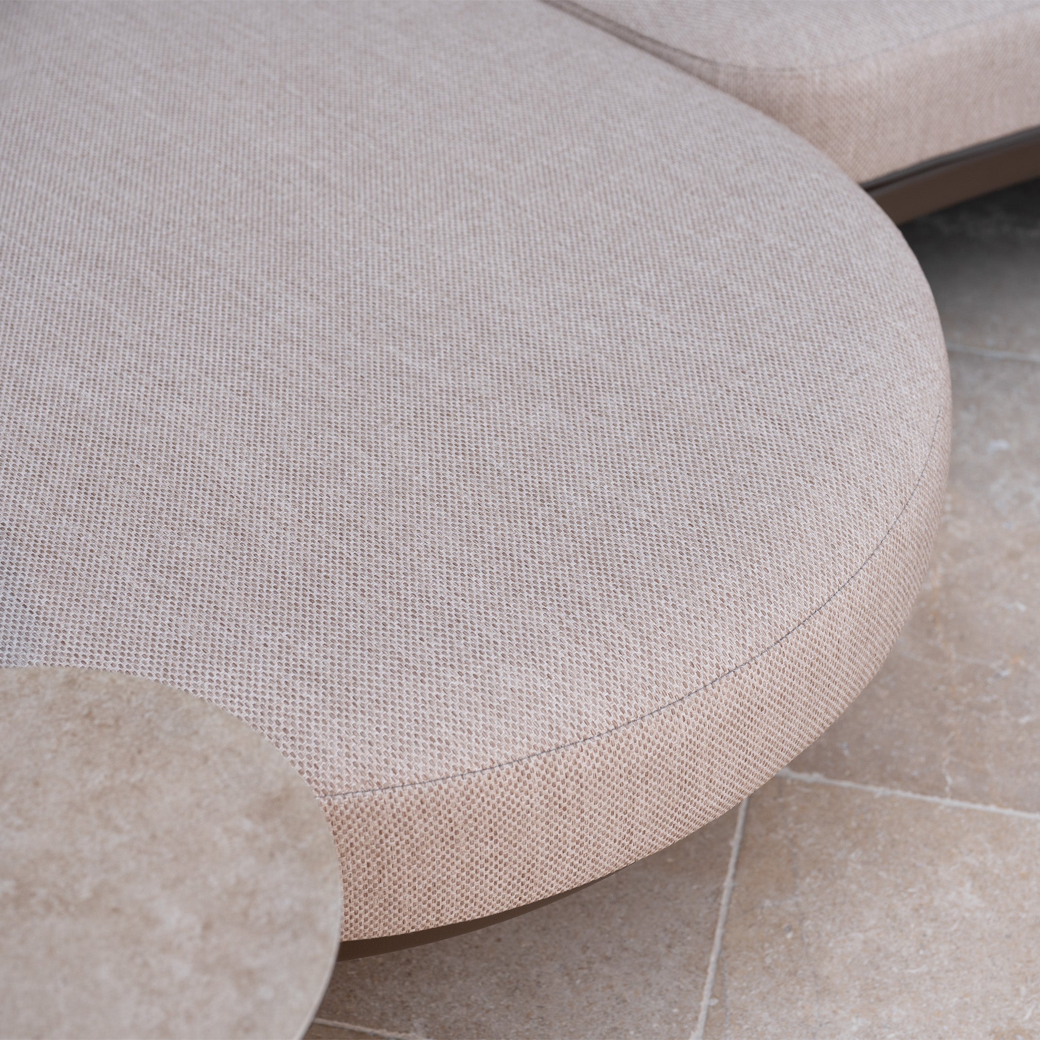 Close-up of the light beige upholstered seat with all-weather cushions from the Sutera Curved Chaise Sofa Set with Armchair in Putty, shown beside a circular stone table on tiled flooring—a stylish choice for outdoor garden furniture.