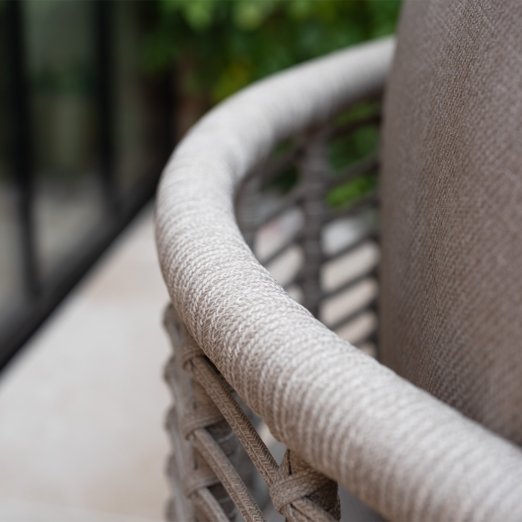Close-up of the Sutera Curved Chaise Sofa Set with Armchair in Putty, highlighting its woven frame, textured armrest, and beige all-weather cushions with blurred greenery in the background.