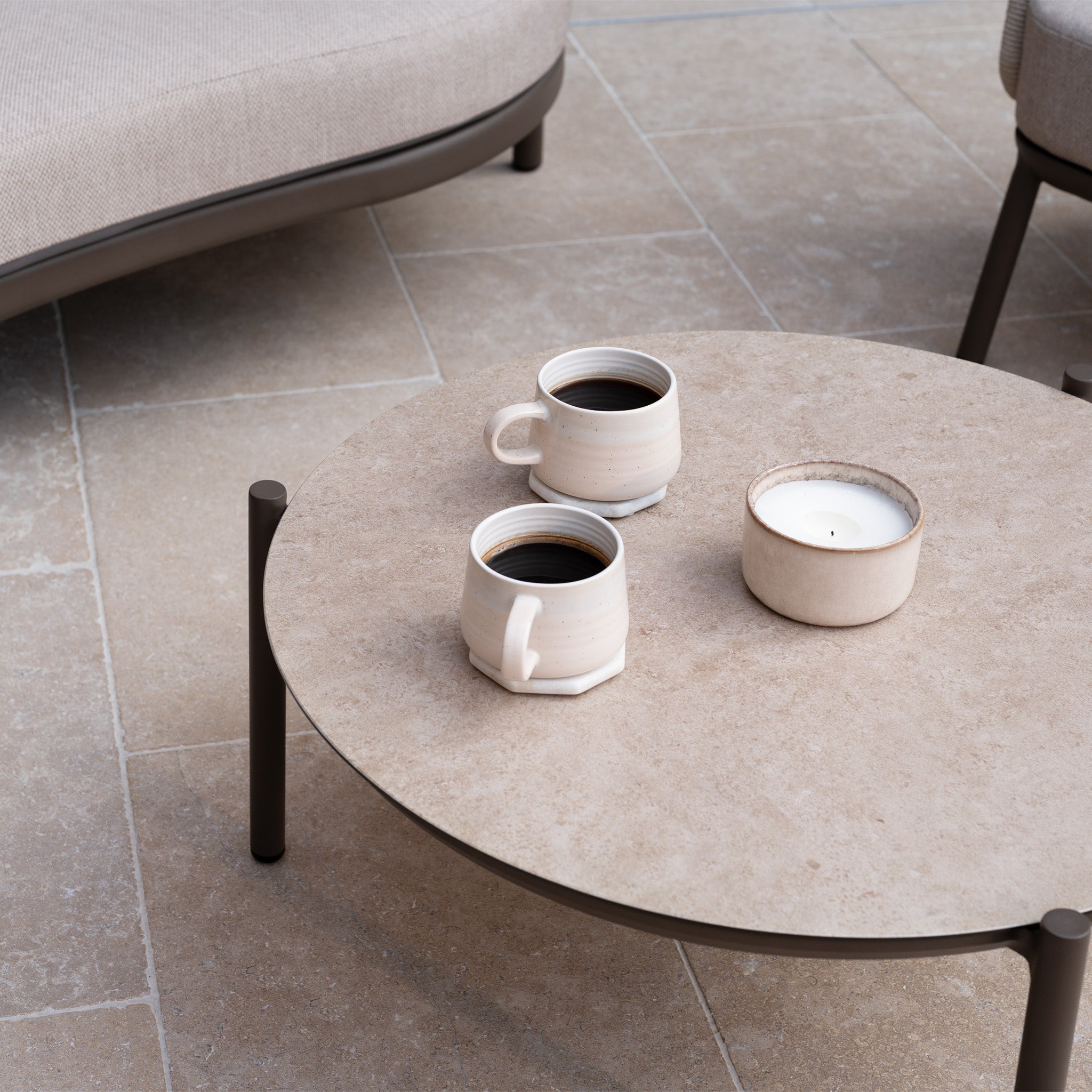 Round coffee table with two mugs and a candle on a tiled patio, paired with outdoor garden furniture and the Sutera Curved Chaise Sofa Set with Armchair in Putty, featuring all-weather cushions for year-round comfort.