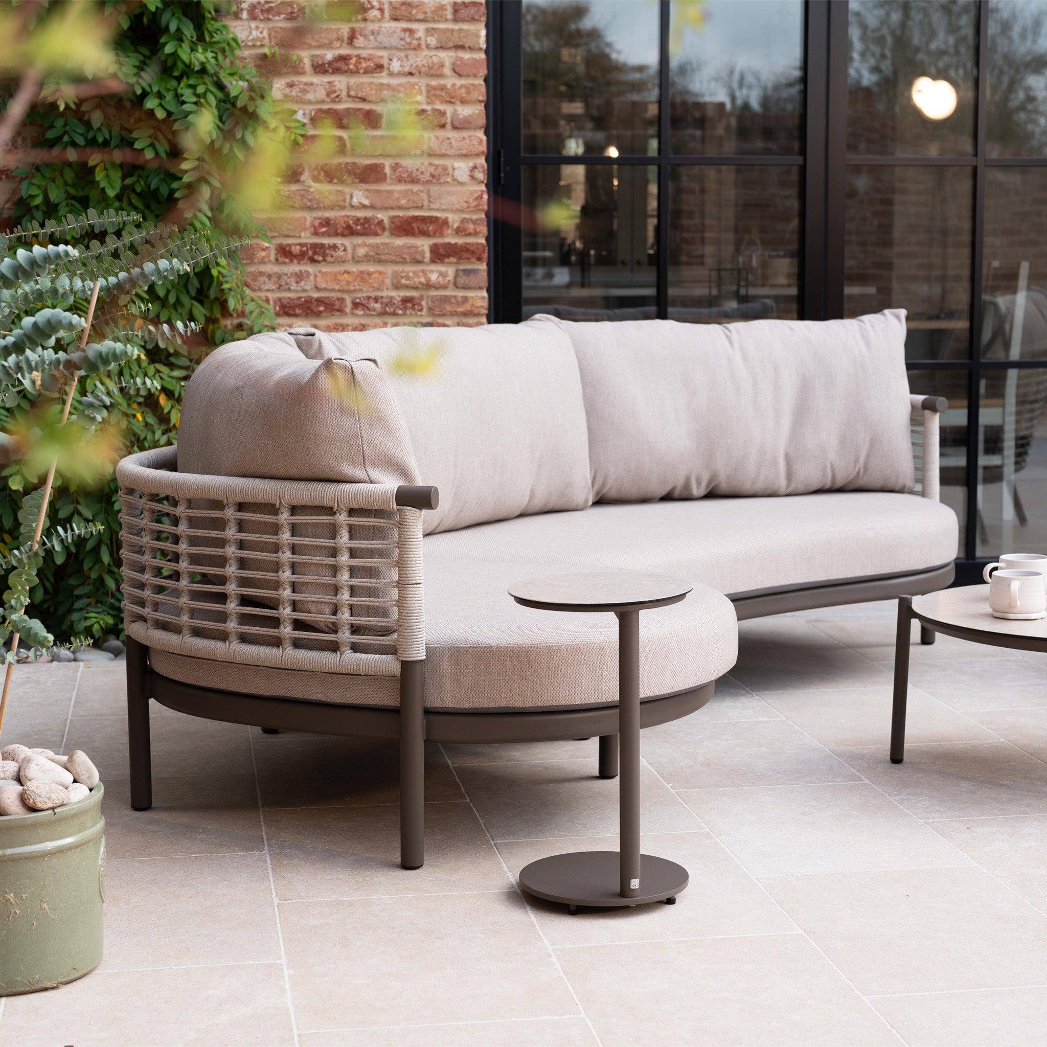 Sutera Curved Chaise Sofa Set with Armchair in Putty, featuring all-weather cushions and pillows, sits on a tiled patio beside small tables and potted plants—ideal outdoor garden furniture for stylish relaxation.