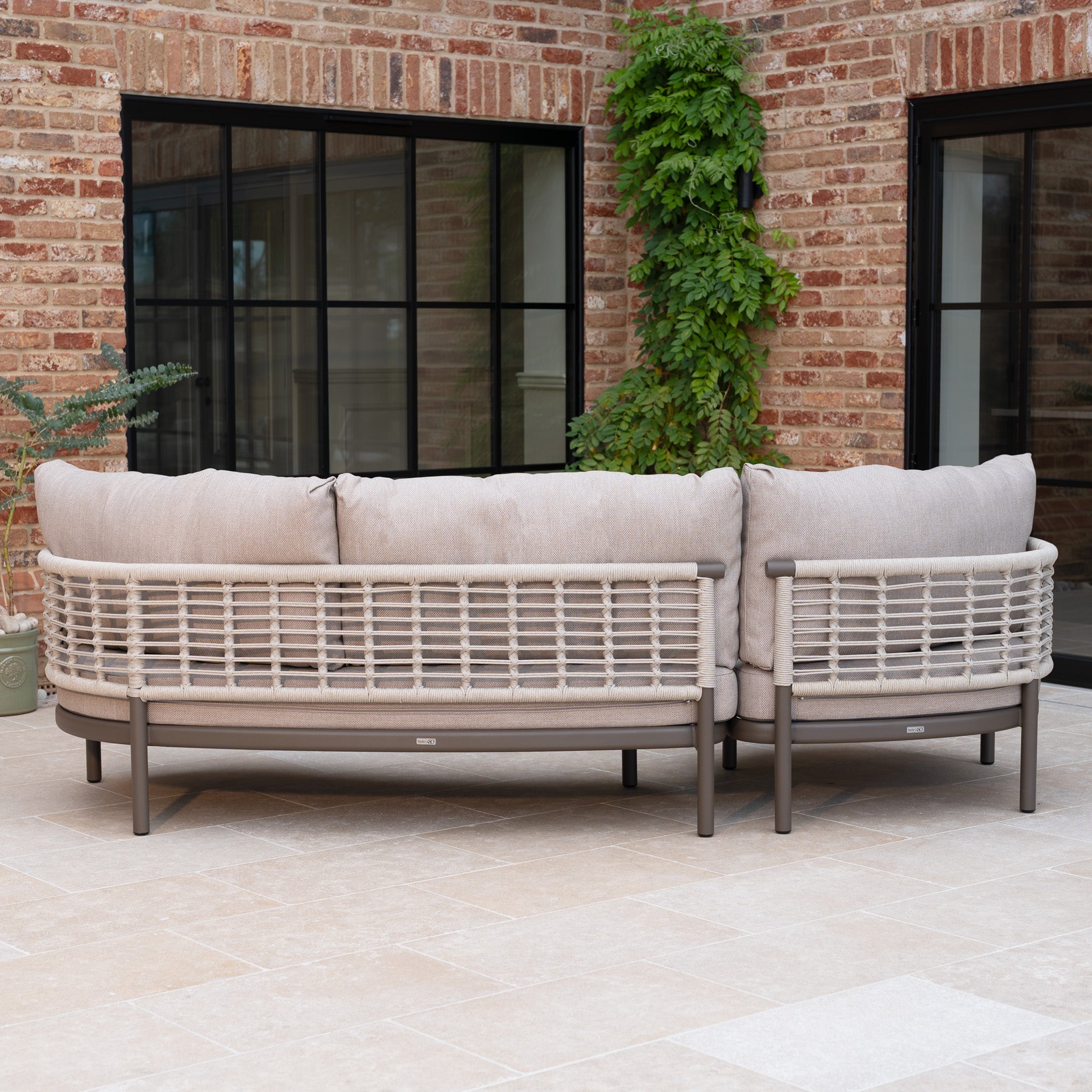 Outdoor patio with a Sutera Curved Chaise Sofa Set with Armchair in Putty, featuring beige all-weather cushions, set against a brick wall and large black-framed windows.