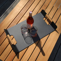 A bottle and two glasses sit on the FREE Tracy Serving Tray with Teak Handles in Charcoal, perfectly placed on a sunlit wooden table. Ideal for enhancing your outdoor space.