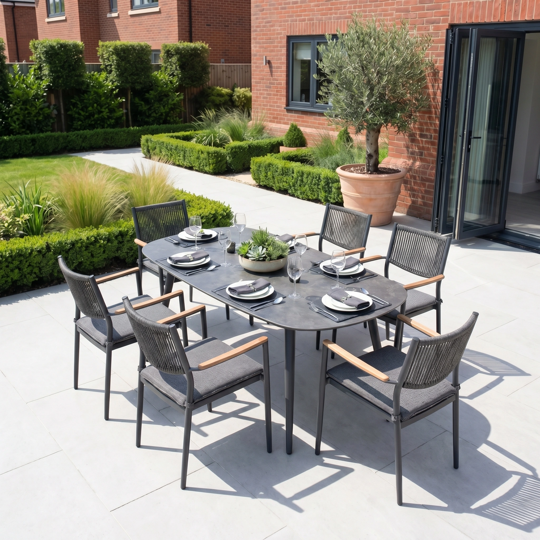 The Tellaro 6 Seat Oval Dining Set in Grey features six chairs on a patio, complemented by lush plants and set against a charming brick house.