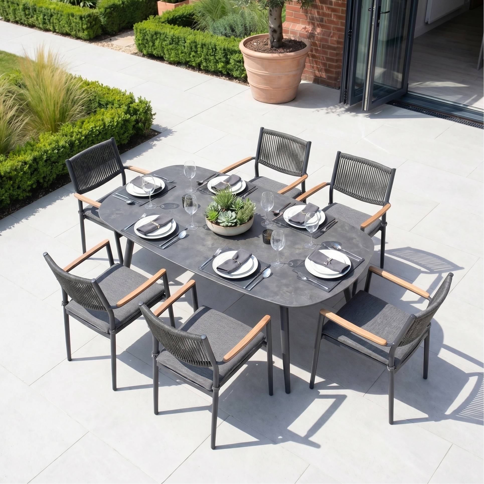 The Tellaro 6 Seat Oval Dining Set in Grey features a modern outdoor dining table for six, complete with plates, glasses, and a succulent centerpiece on a stylish patio.