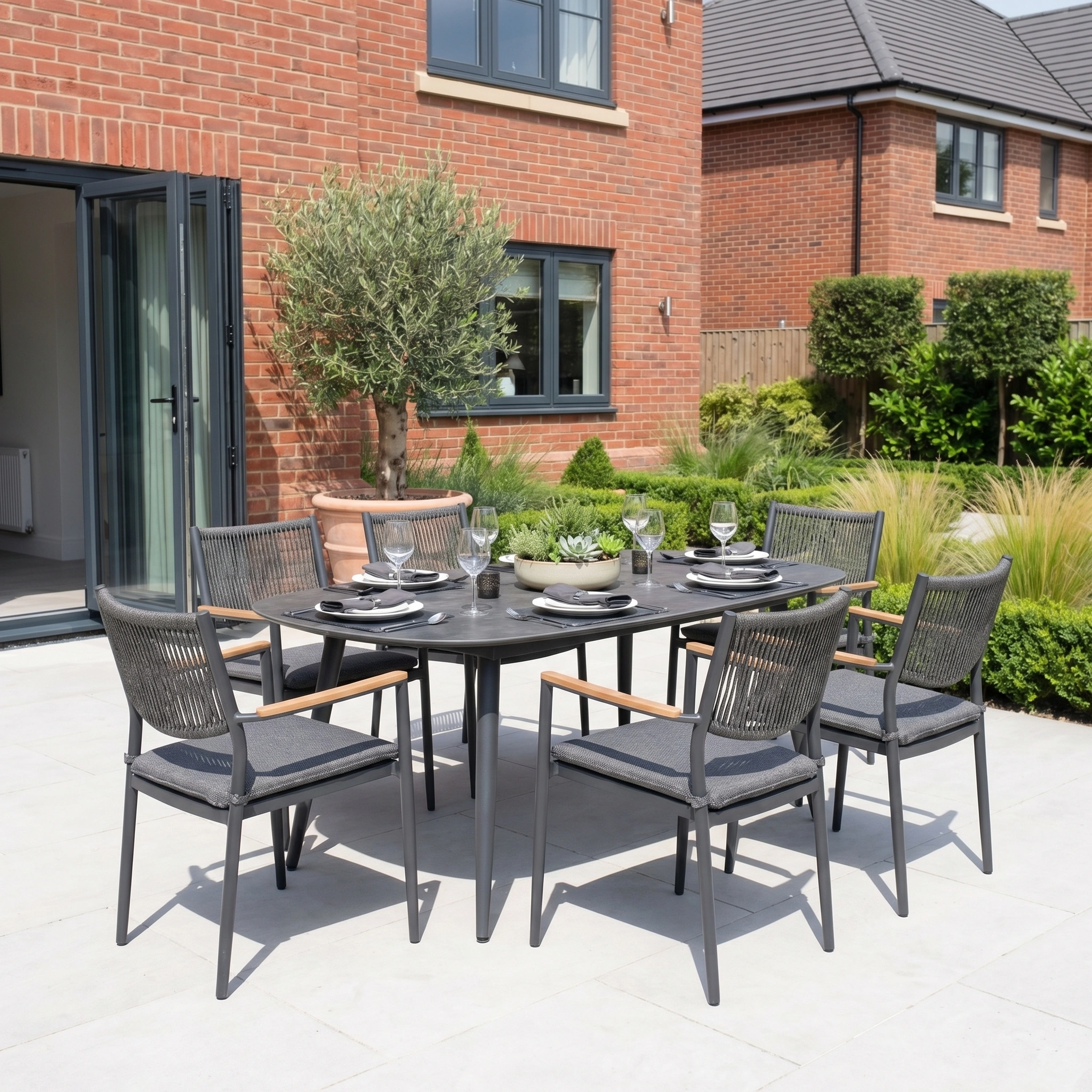 The Tellaro 6 Seat Oval Dining Set in Grey features a modern dining table and six chairs, arranged on a patio before a brick house with a lush garden backdrop, perfect for outdoor meals.