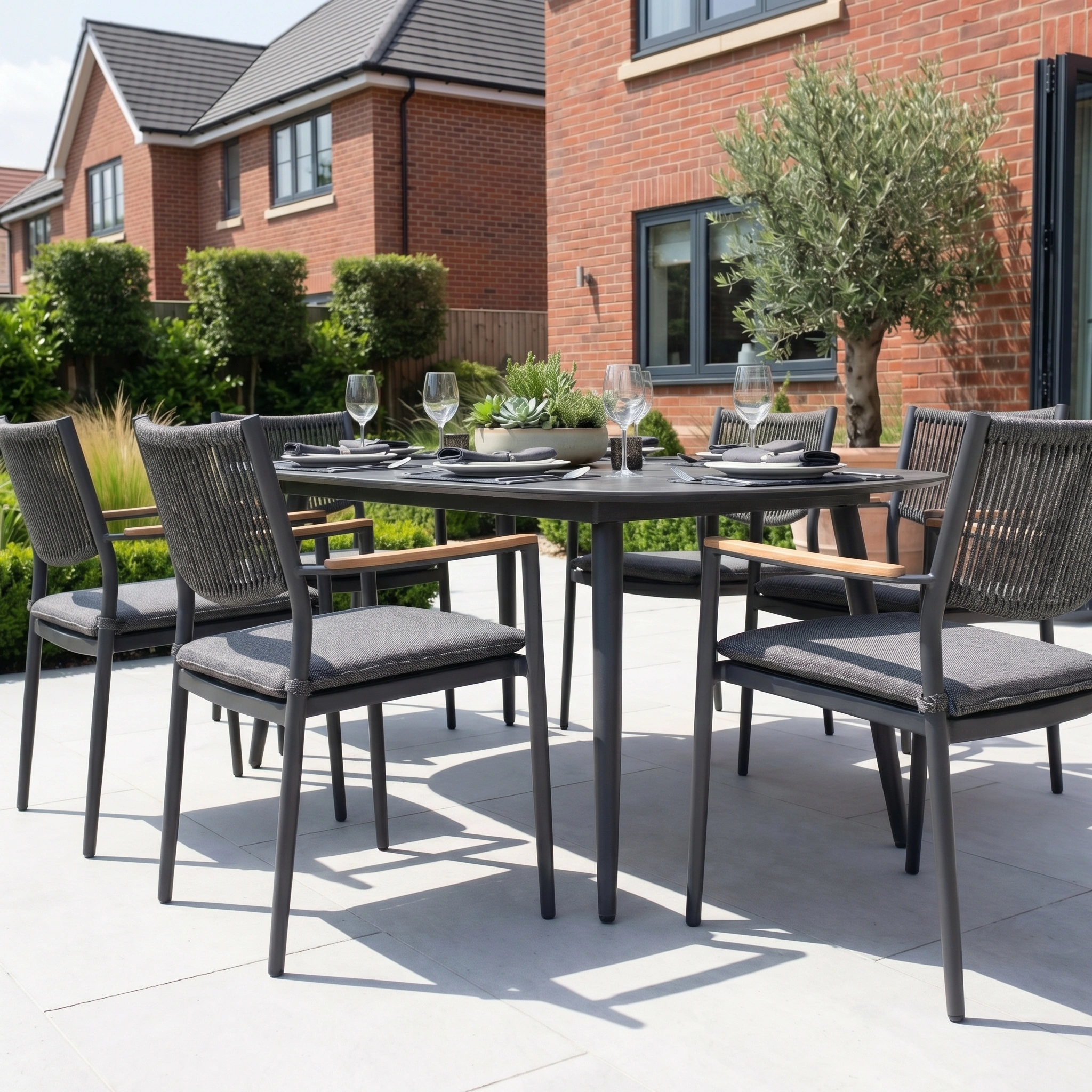 The Tellaro 6 Seat Oval Dining Set in Grey is arranged for a meal on a patio beside a brick house, blending comfort and style in modern garden furniture.