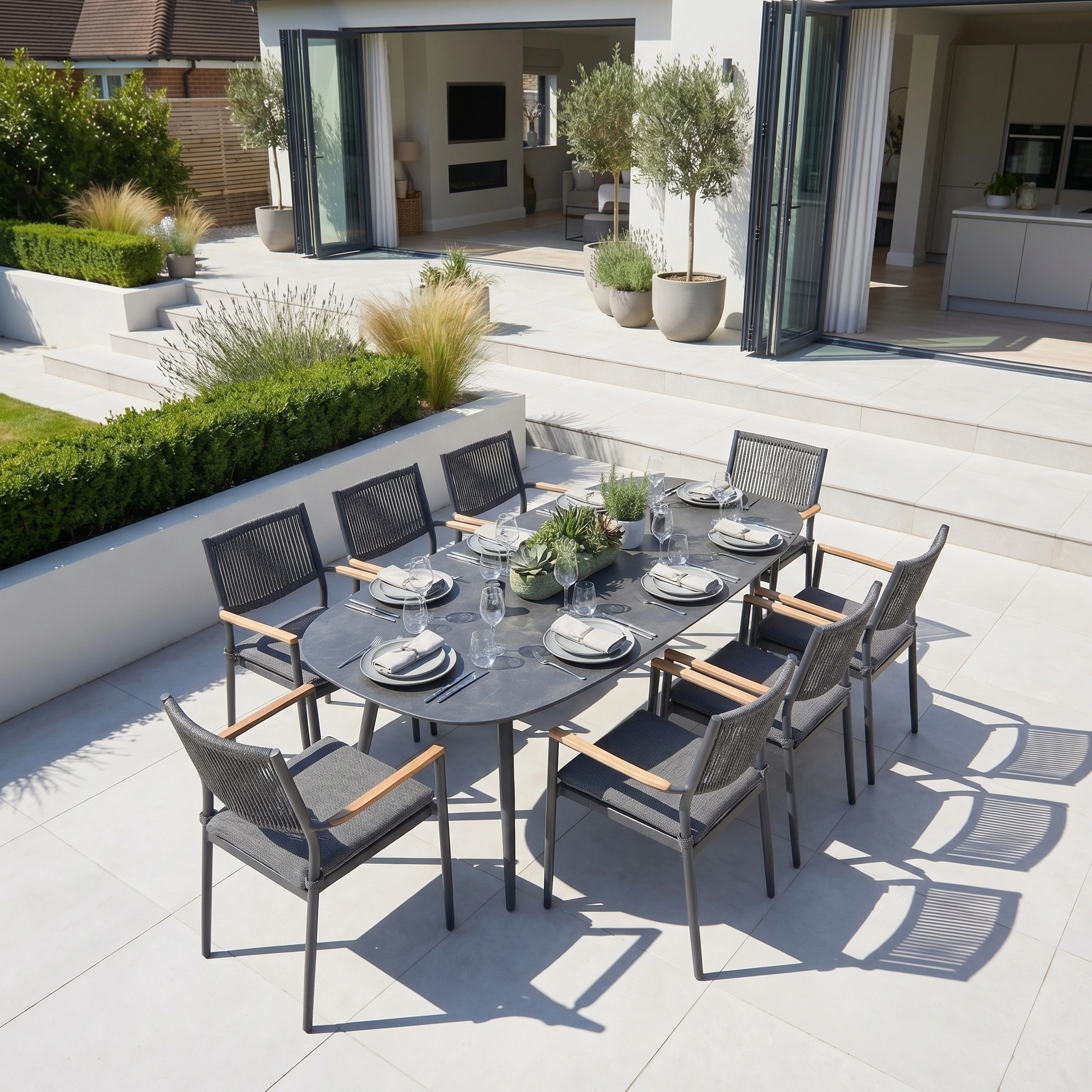 The Tellaro 8 Seat Oval Dining Set in Grey features an elegant table and dark grey chairs, perfect for outdoor dining on a white tiled patio with lush green landscaping.