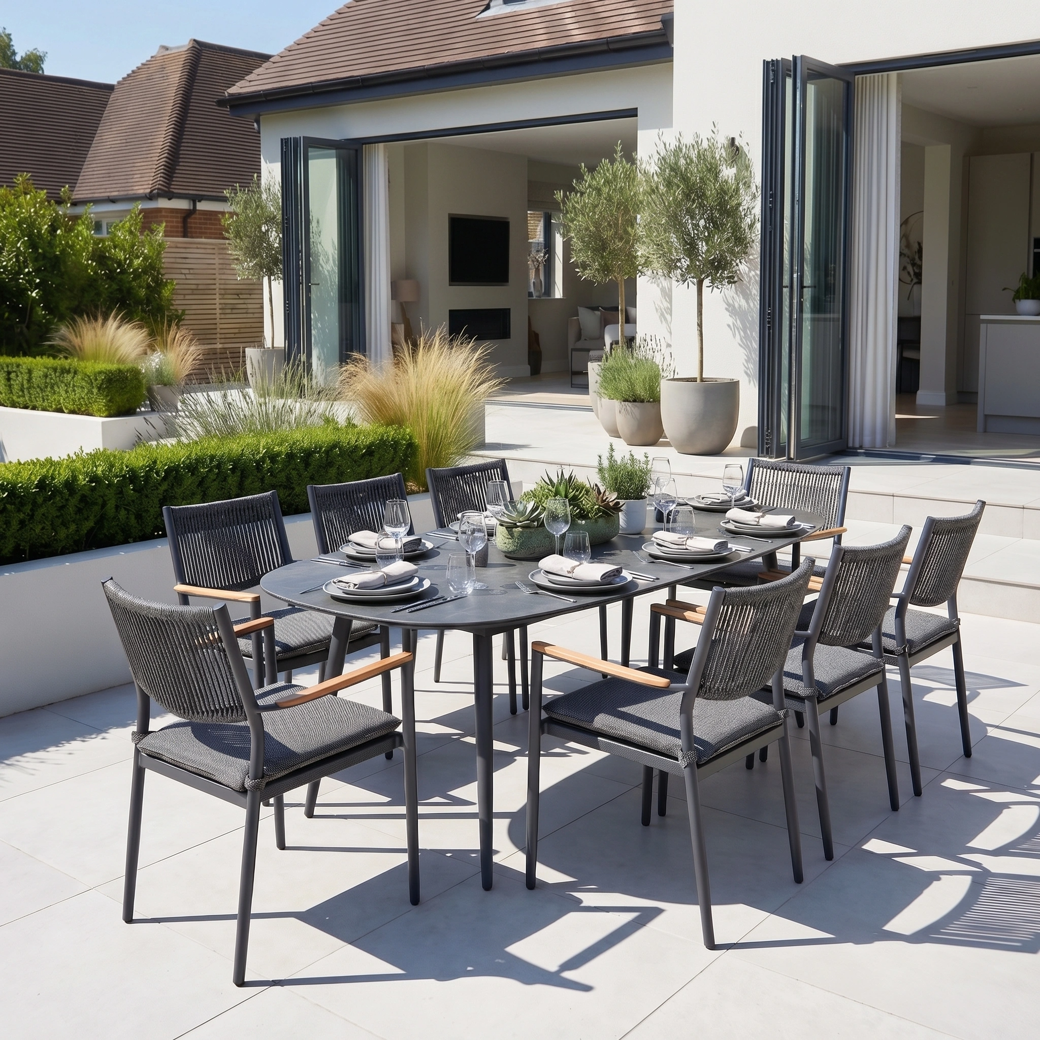 The Tellaro 8 Seat Oval Dining Set in Grey features eight chairs and a spacious table with a sleek grey aluminum finish, ideal for outdoor dining on a sunny patio beside a modern home.