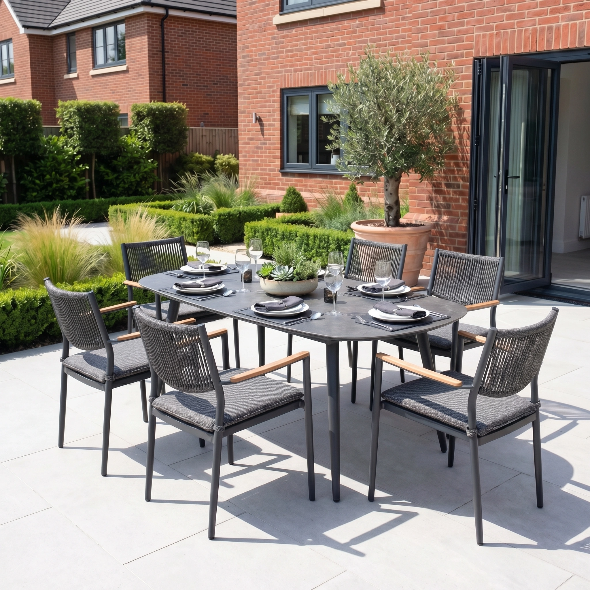 The Tellaro 8 Seat Oval Dining Set in Grey offers modern outdoor dining for eight, featuring sleek grey aluminium on a patio with lush greenery and brick houses in the background.