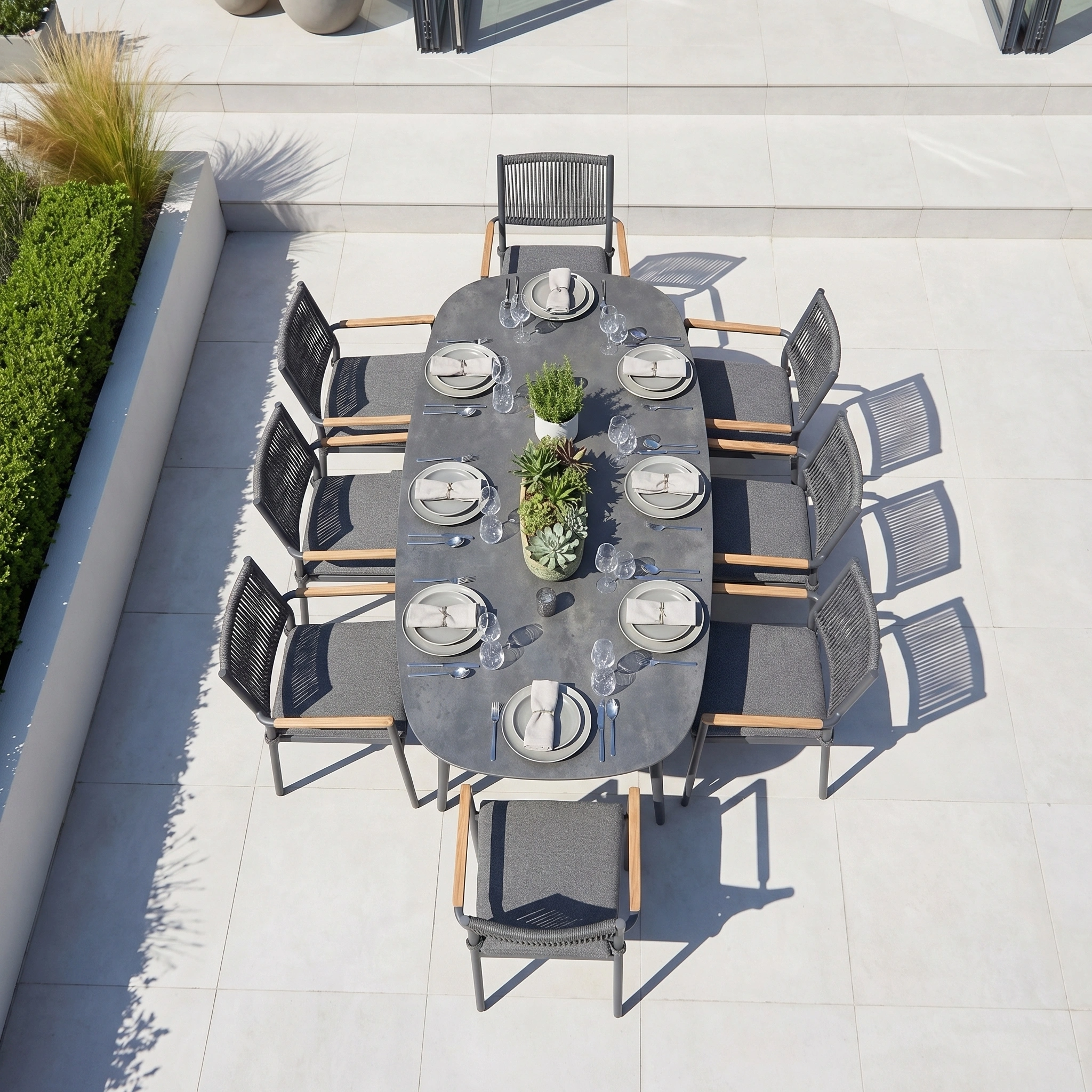 The Tellaro 8 Seat Oval Dining Set in Grey features sleek gray chairs and tabletop plants on a tiled patio—ideal for stylish outdoor dining.