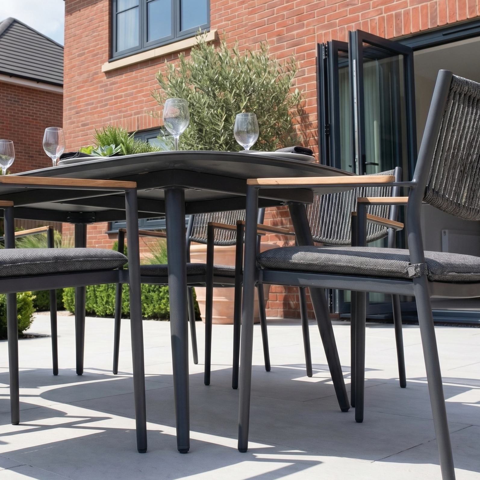 The Tellaro 6 Seat Oval Dining Set in Grey is arranged on a patio beside a brick house with open glass doors, offering modern garden furniture for outdoor dining.