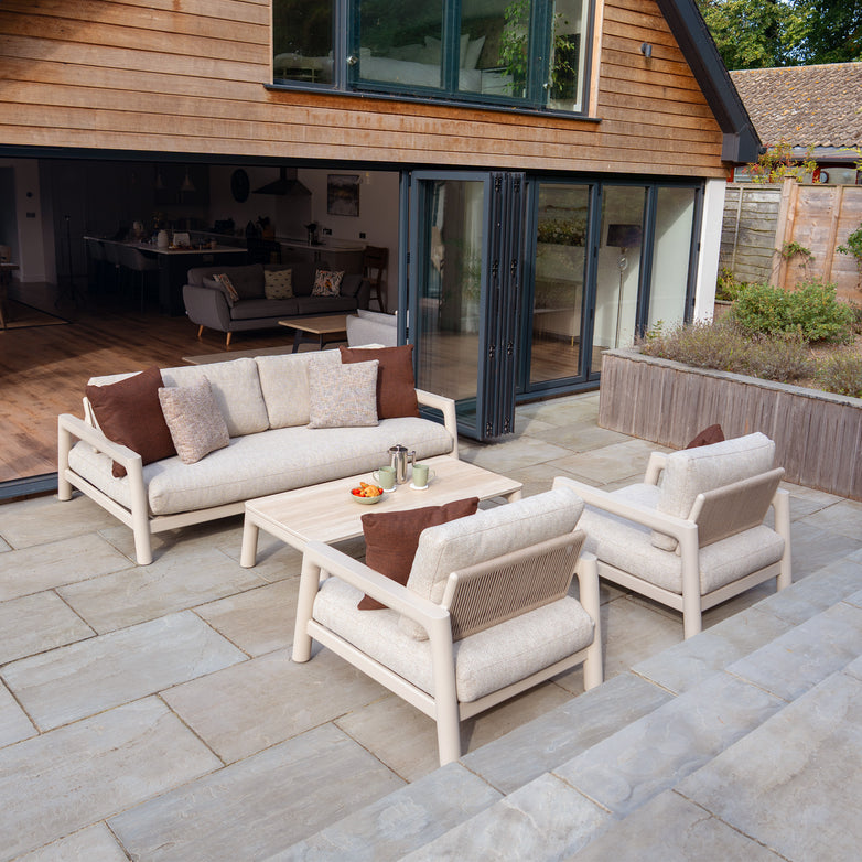 Outdoor patio with the Theia 3 Seat Sofa Set in Oatmeal, a coffee table, and sliding glass doors opening to a modern home.