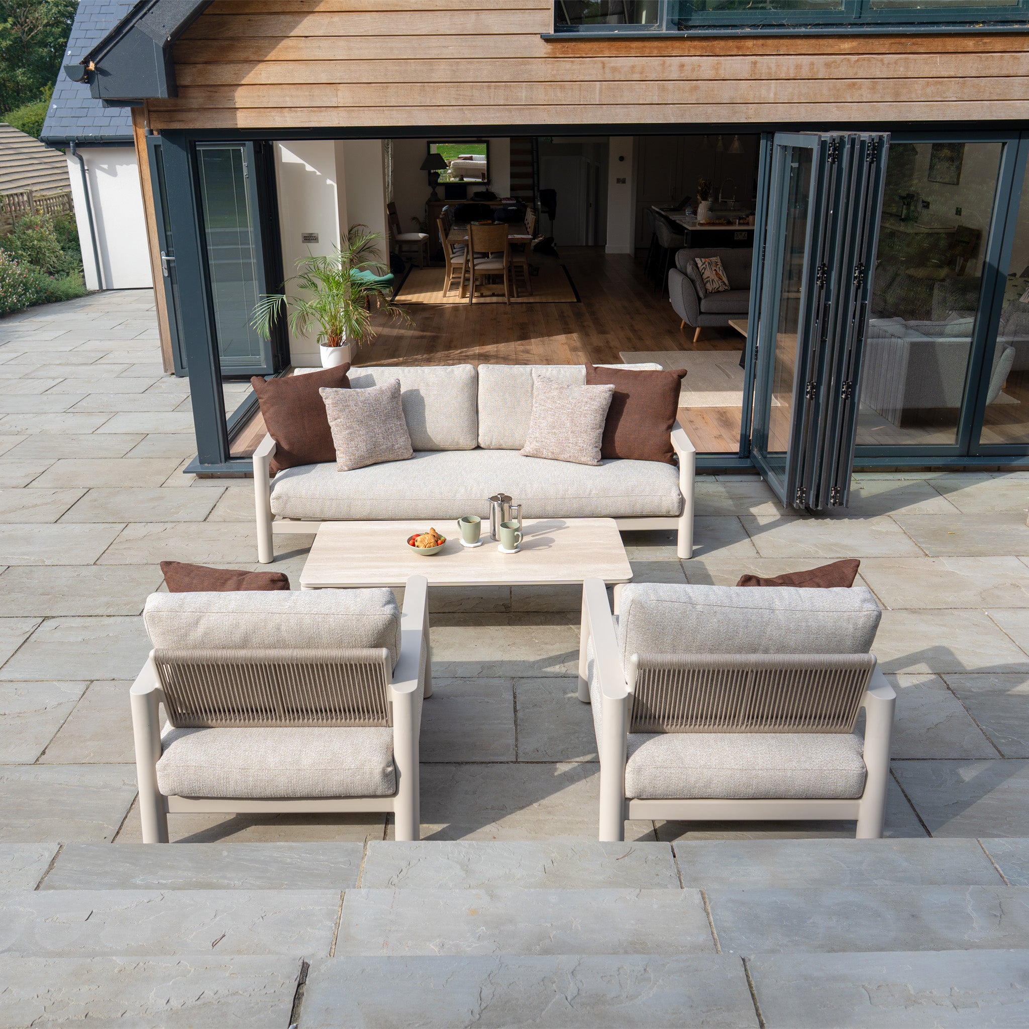 Modern outdoor patio featuring the Theia 3 Seat Sofa Set in Oatmeal with plush cushions, a stylish coffee table, and open doors connecting to the living area.