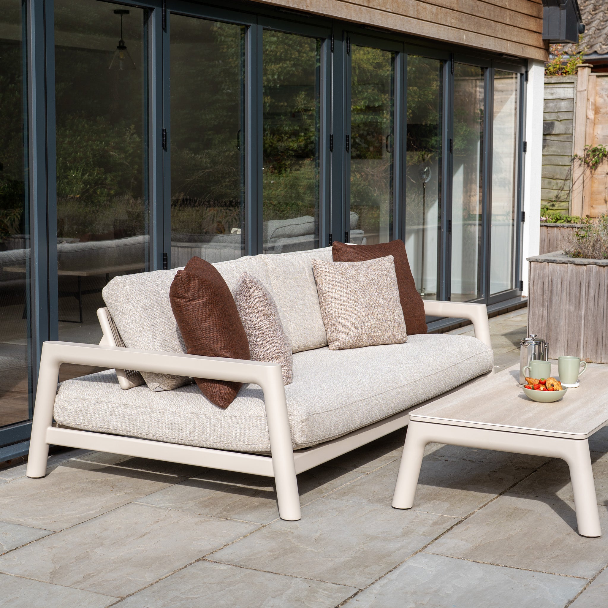 The outdoor patio features the Theia 3 Seat Sofa Set in Oatmeal with brown cushions and a matching table holding tea, cups, and a bowl of fruit.