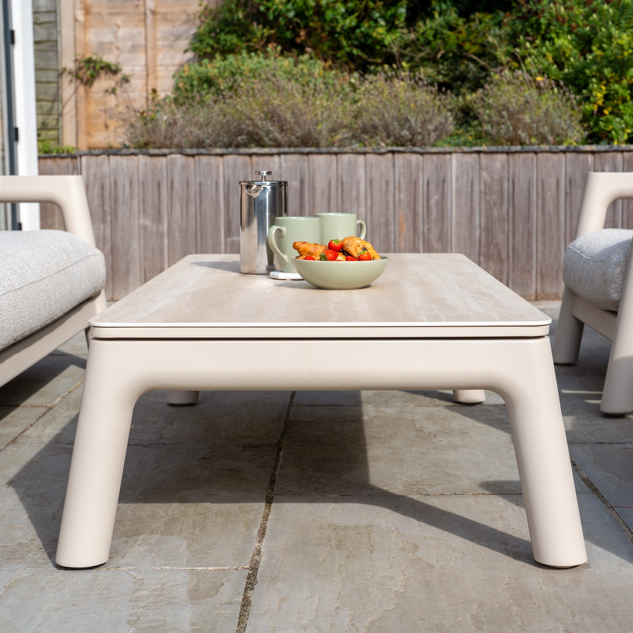 The Theia 3 Seat Sofa Set in Oatmeal features a beige outdoor table with mugs, a French press, and a bowl of strawberries, flanked by cushioned chairs and complemented by stylish modular garden furniture.
