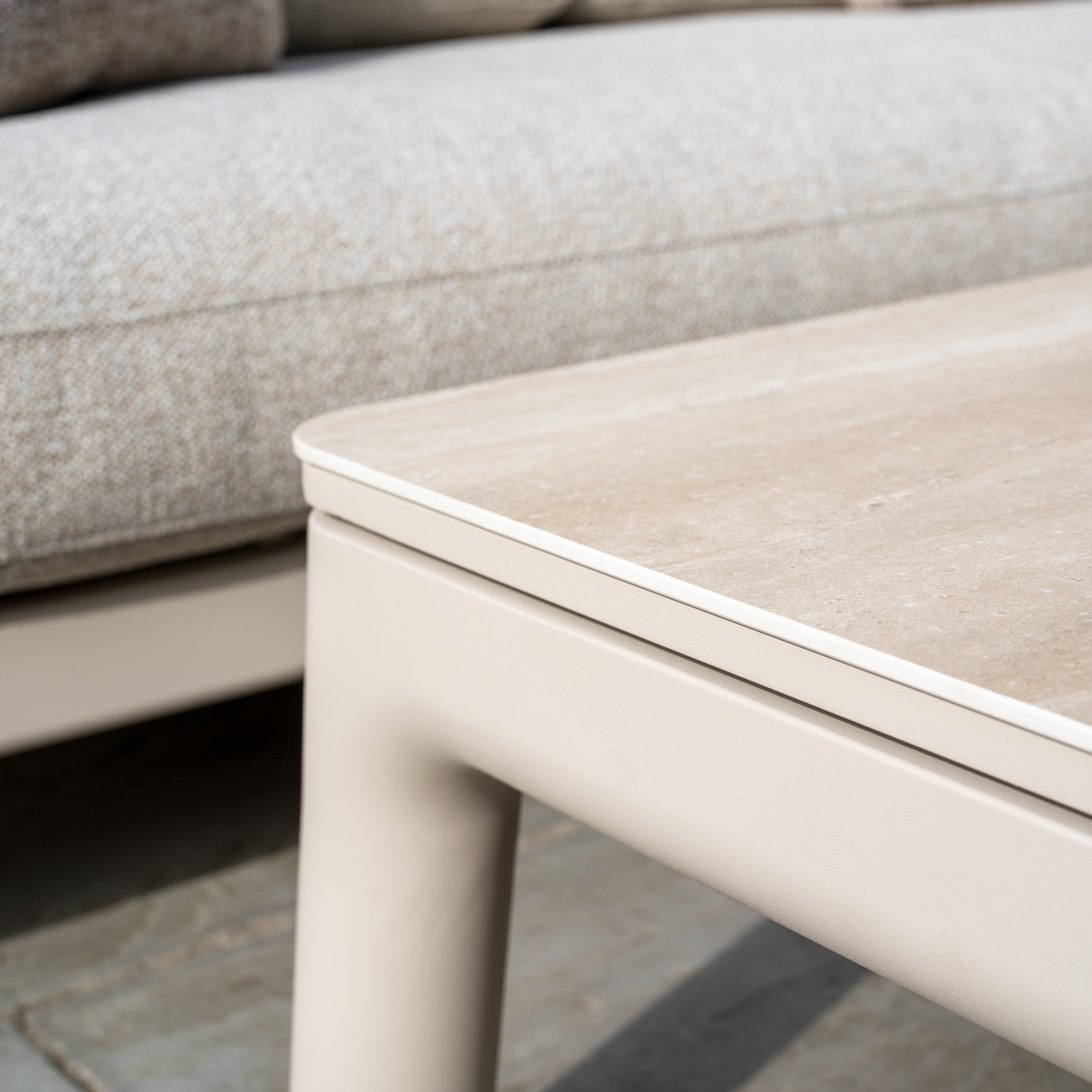 Close-up of a beige outdoor coffee table beside Theia 3 Seat Sofa Set in Oatmeal with light gray cushions, placed on a stone patio.