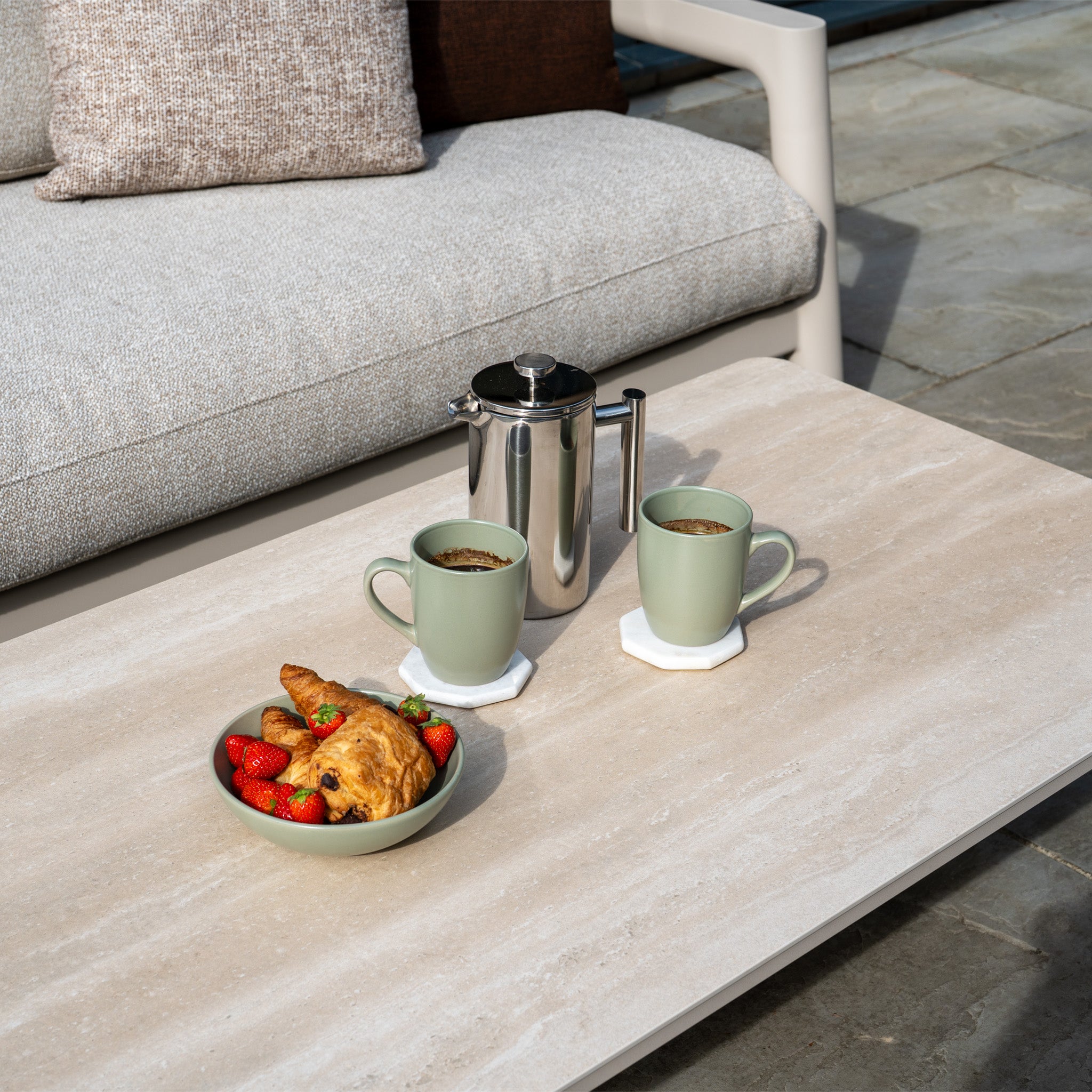 Two mugs of coffee, a bowl of strawberries, and a croissant on a patio table in front of the Theia 3 Seat Sofa Set in Oatmeal.