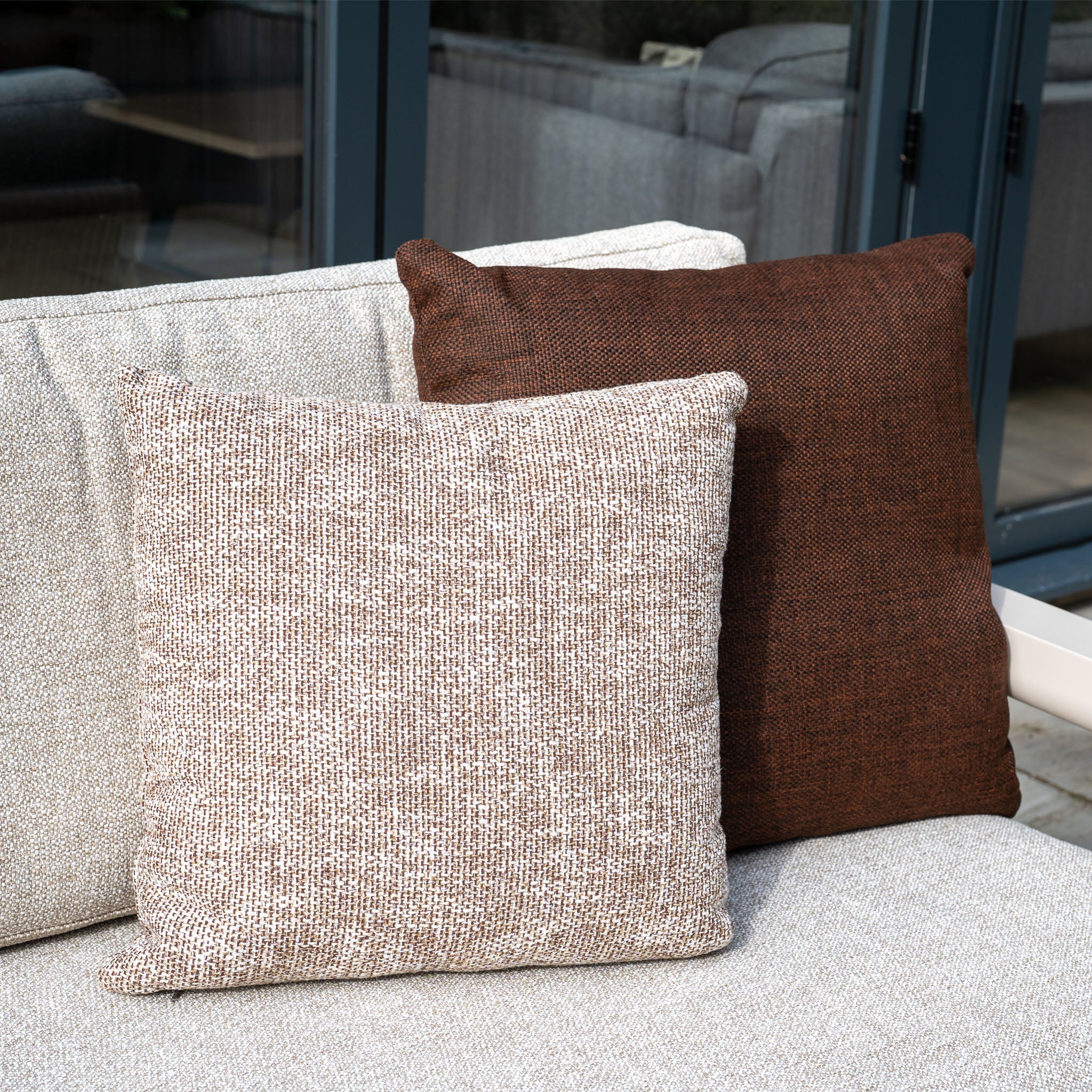Two square cushions, one beige and one brown, rest on a light Theia 3 Seat Sofa Set in Oatmeal—a sleek modular garden piece—positioned near glass doors.