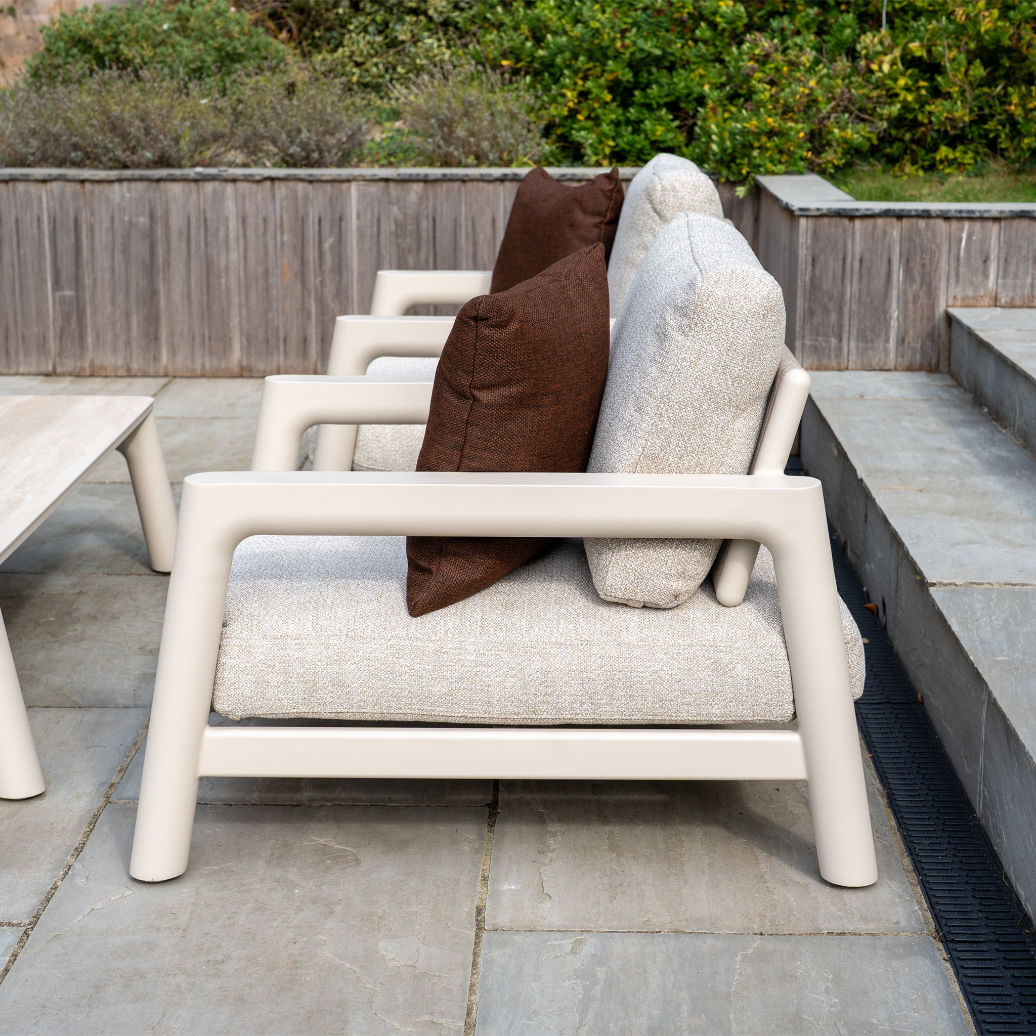 Side view of three beige outdoor armchairs with brown cushions on a stone patio near greenery—perfect for pairing with modular garden furniture or an outdoor sofa like the Theia 3 Seat Sofa Set in Oatmeal.