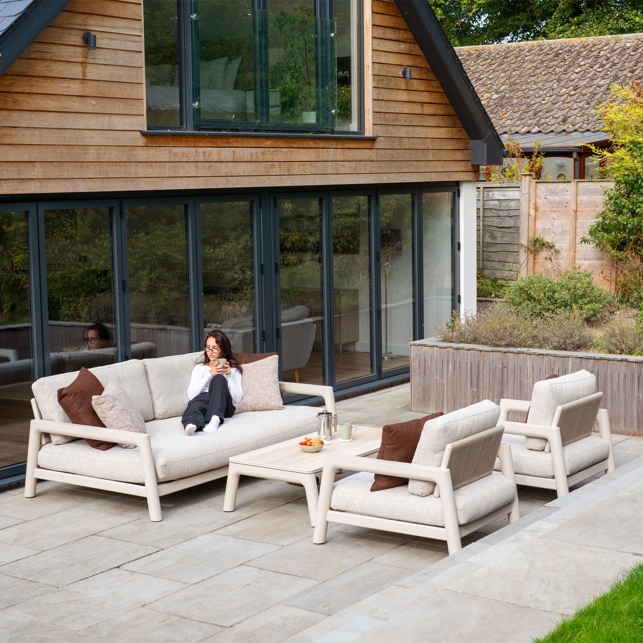 A woman relaxes on the Theia 3 Seat Sofa Set in Oatmeal, using her phone, with sleek patio furniture and a modern house in the background.