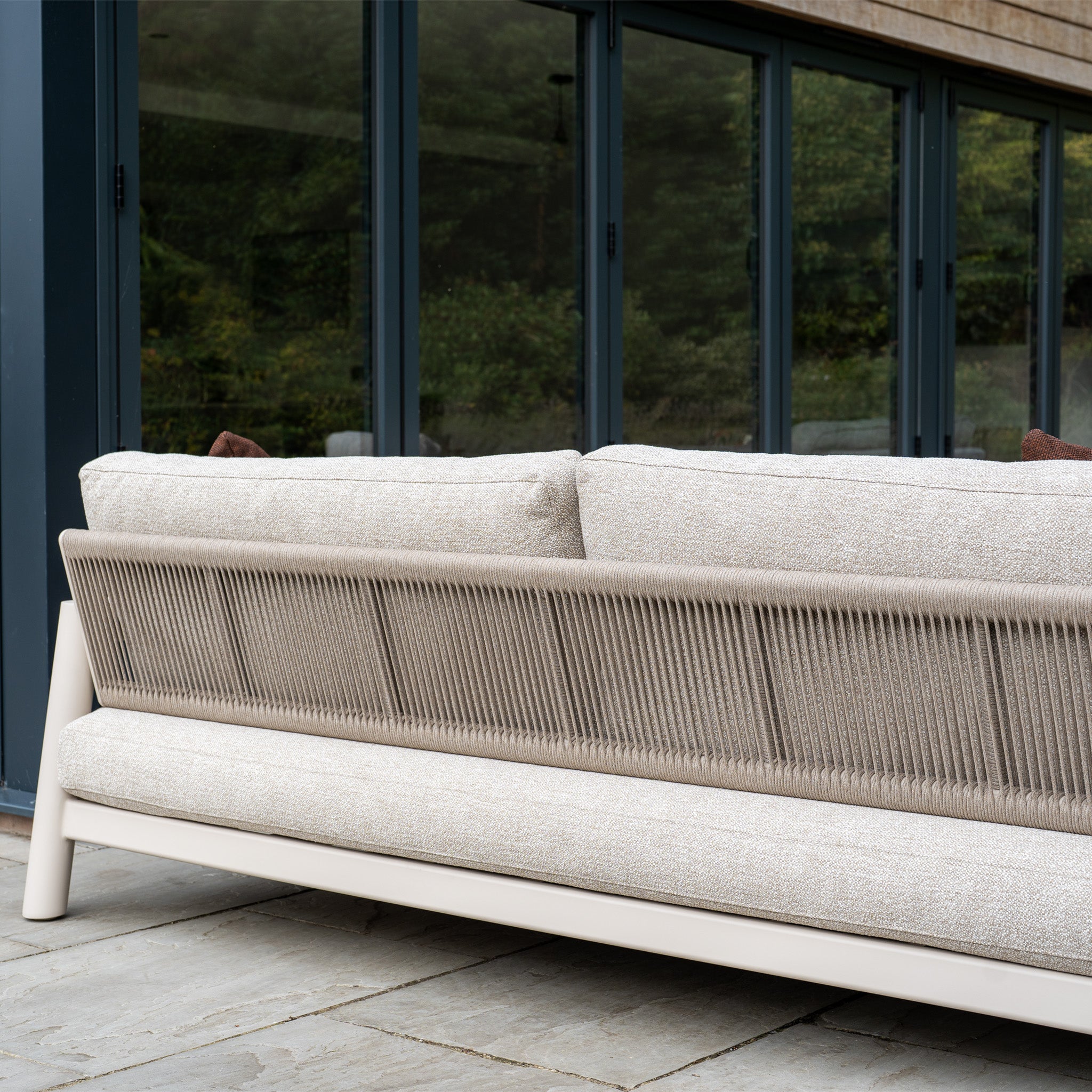 The Theia 3 Seat Sofa Set in Oatmeal, with its woven backrest, brings modern elegance to a stone patio by glass doors—an ideal choice for stylish modular garden furniture.