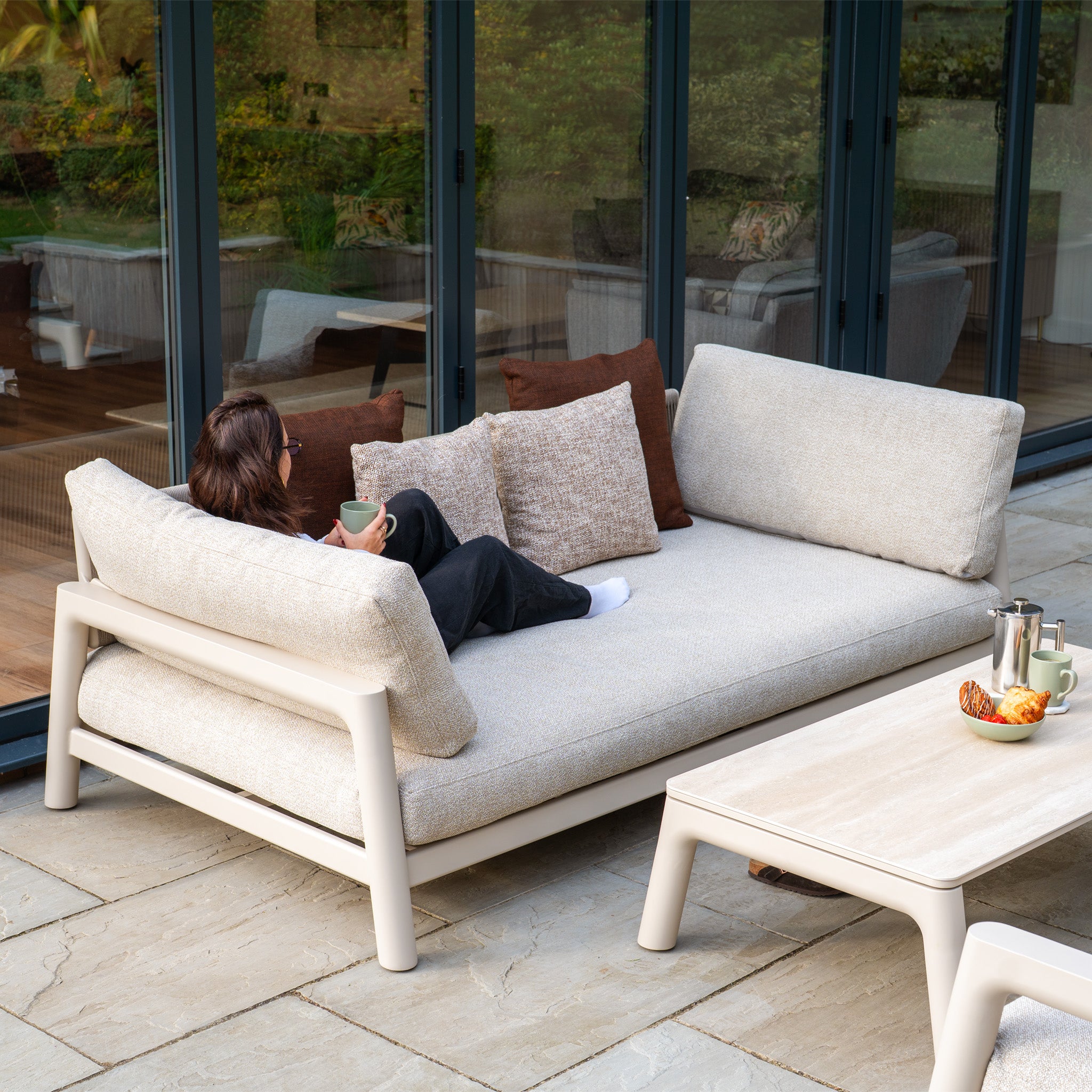 A woman relaxes on the Theia 3 Seat Sofa Set in Oatmeal with pillows, holding a mug beside a breakfast table—showcasing the inviting comfort of this modular garden furniture.