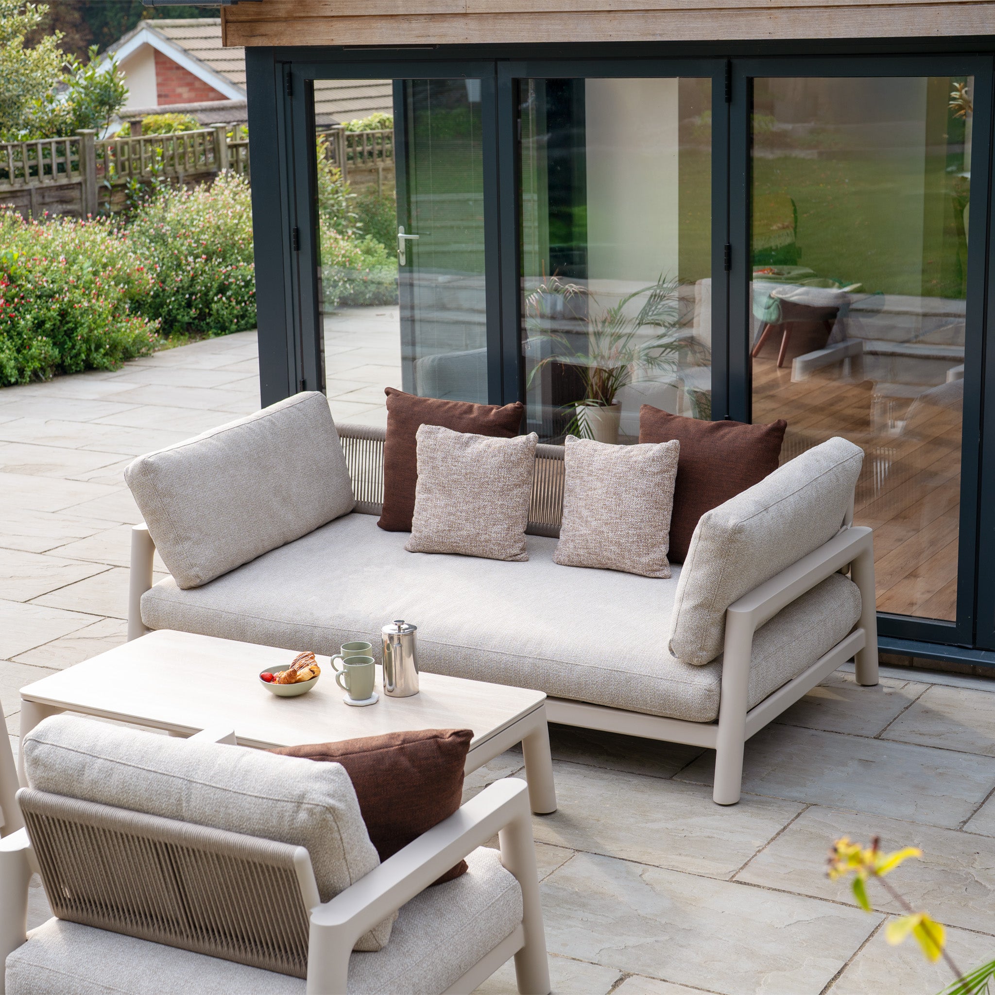 The Theia 3 Seat Sofa Set in Oatmeal, featuring brown cushions and a matching table with snacks and drinks, sits on a stone patio beside glass doors.