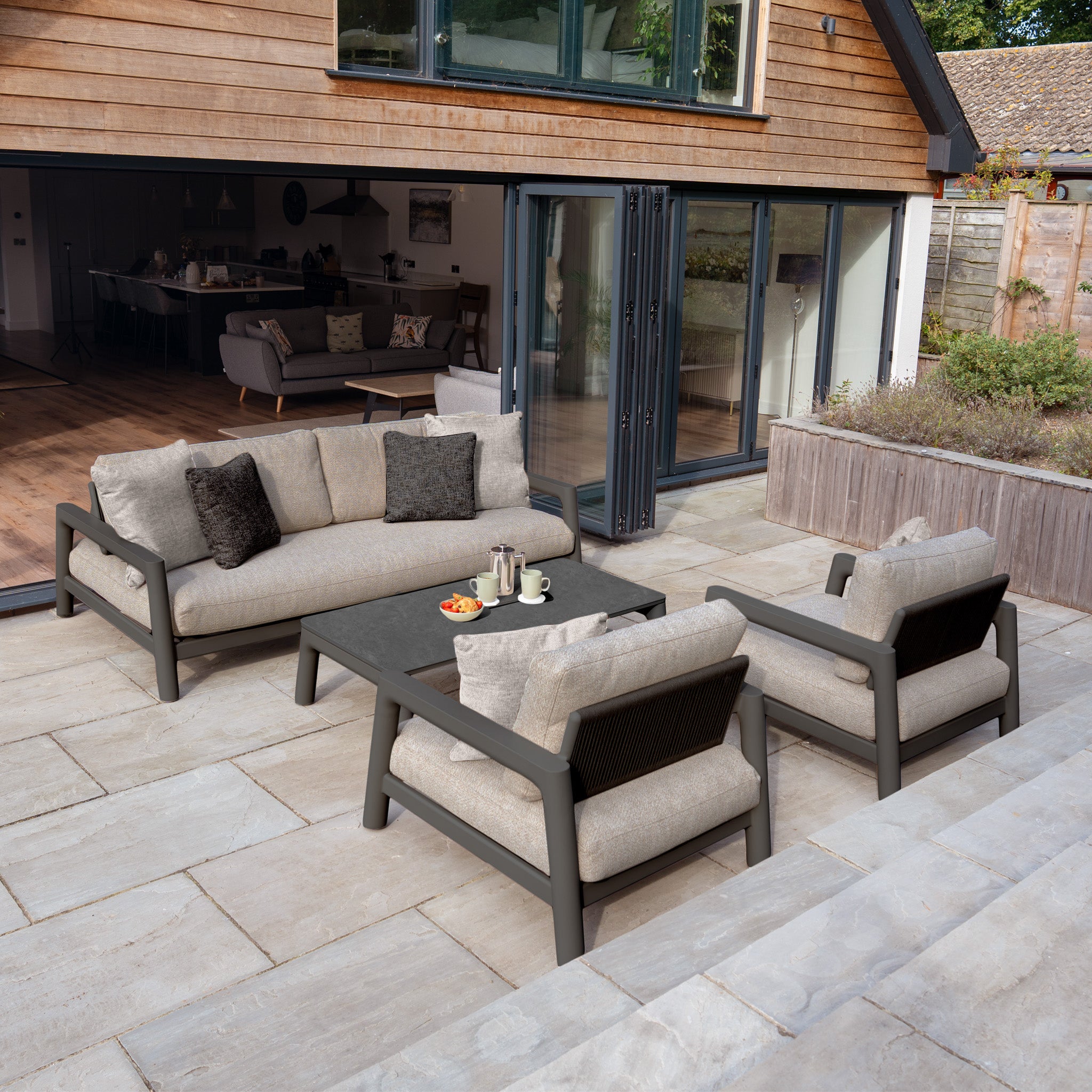 Modern patio featuring the Theia 3 Seat Sofa Set in Truffle with beige cushions, two armchairs, and a coffee table set for tea, creating a stylish outdoor arrangement.