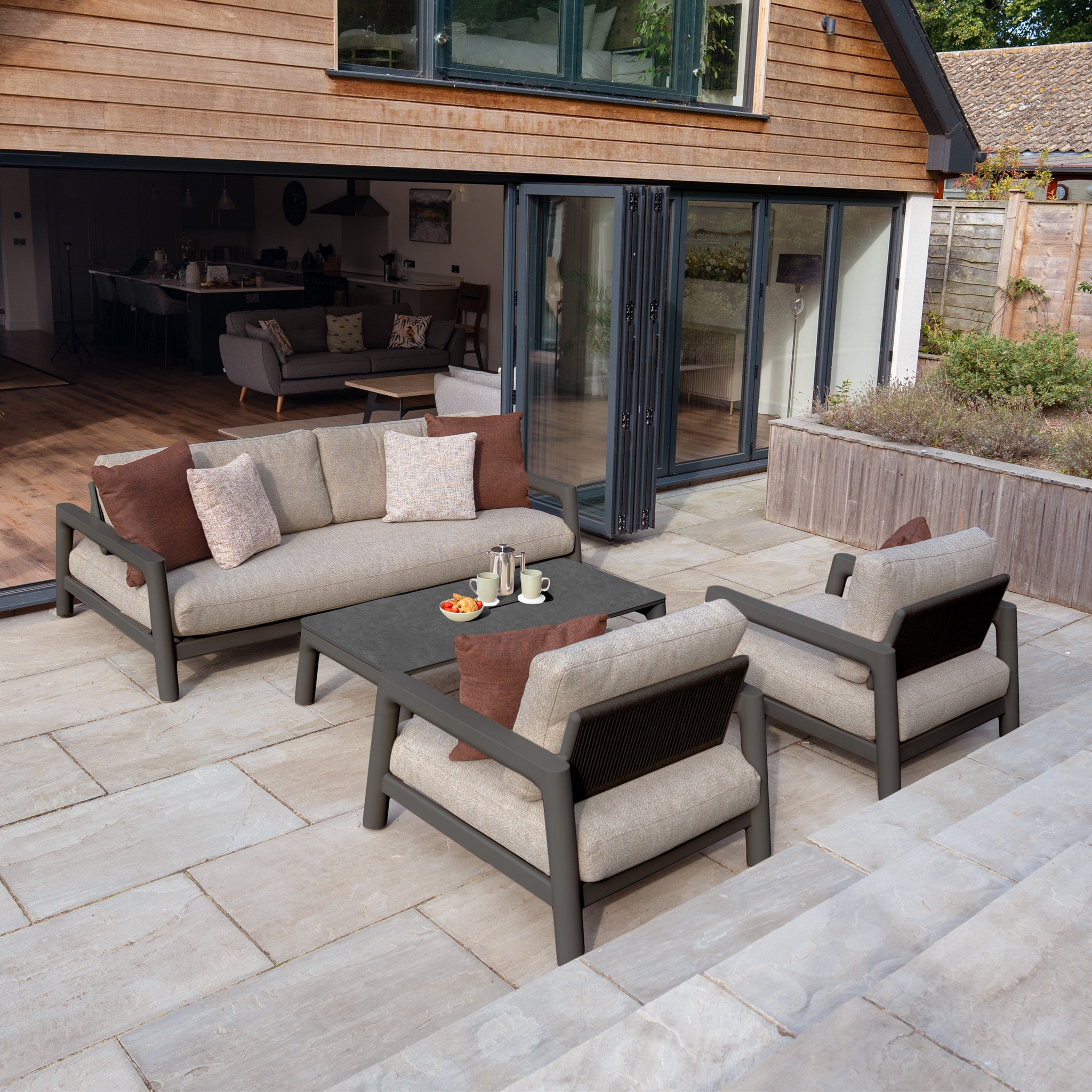 Contemporary patio showcases the Theia 3 Seat Sofa Set in Truffle—aluminium sofa, two chairs, coffee table, and cushions—arranged outside a home with open glass doors.