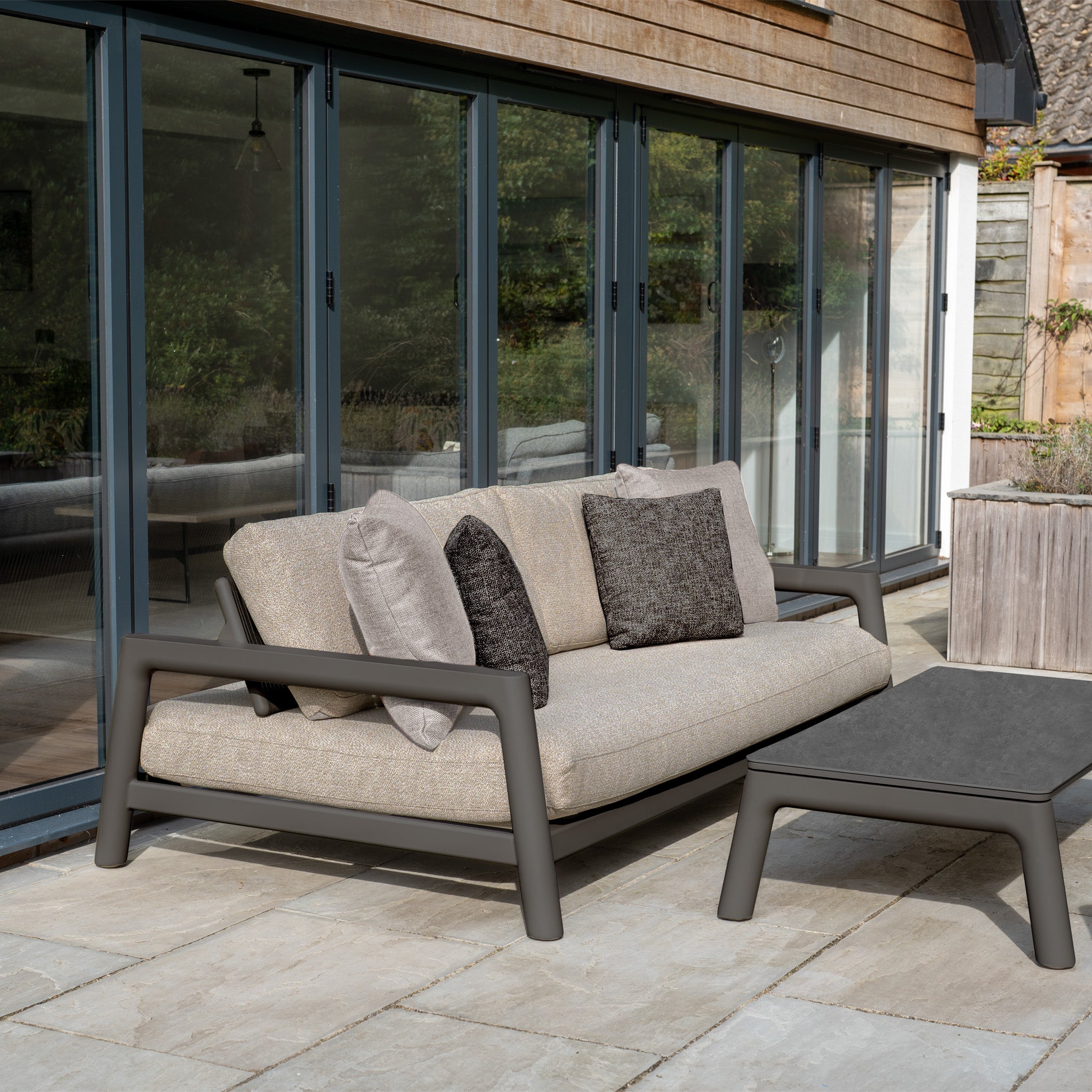 The Theia 3 Seat Sofa Set in Truffle features neutral cushions and a coffee table, arranged on a tiled patio by glass doors.