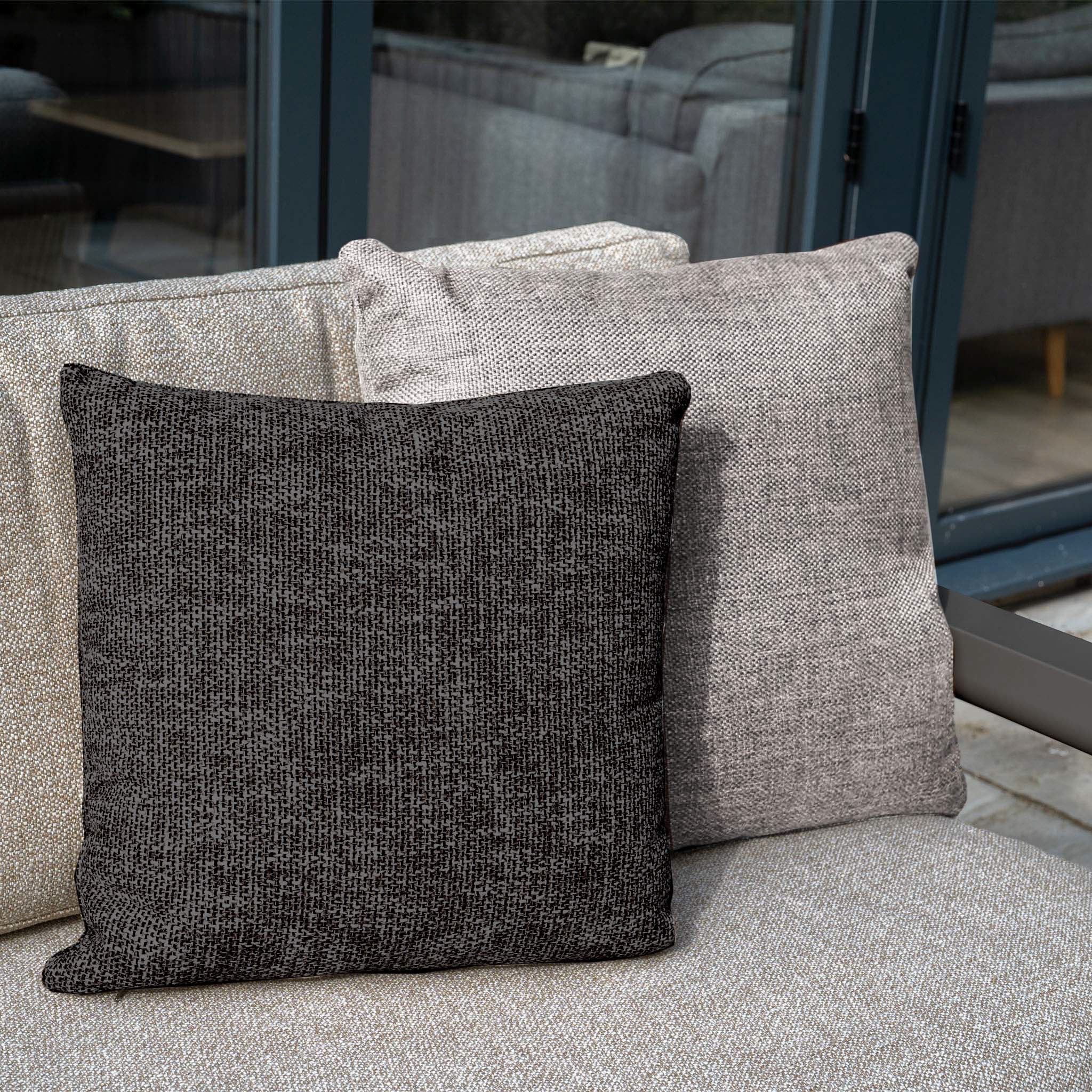 Two textured throw pillows, one dark gray and one light gray, rest on Theia 3 Seat Sofa Set in Truffle—a stylish modular outdoor piece placed near glass doors.