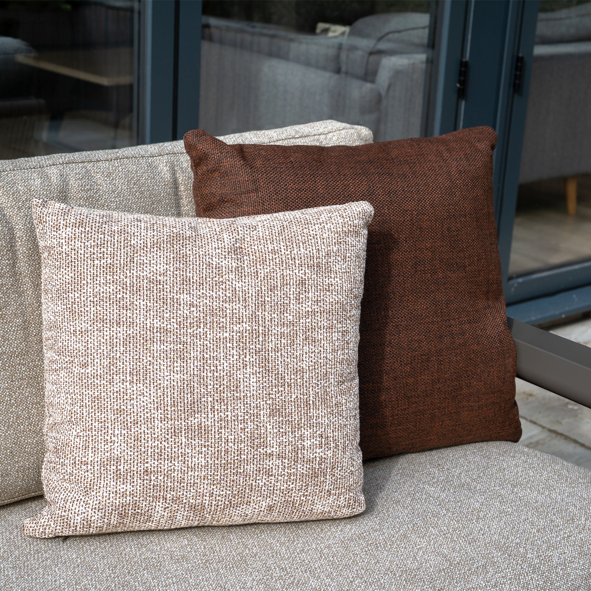 Two textured square cushions—one light beige, one dark brown—adorn the Theia 3 Seat Sofa Set in Truffle near glass doors, adding an inviting touch to your outdoor furniture arrangement.