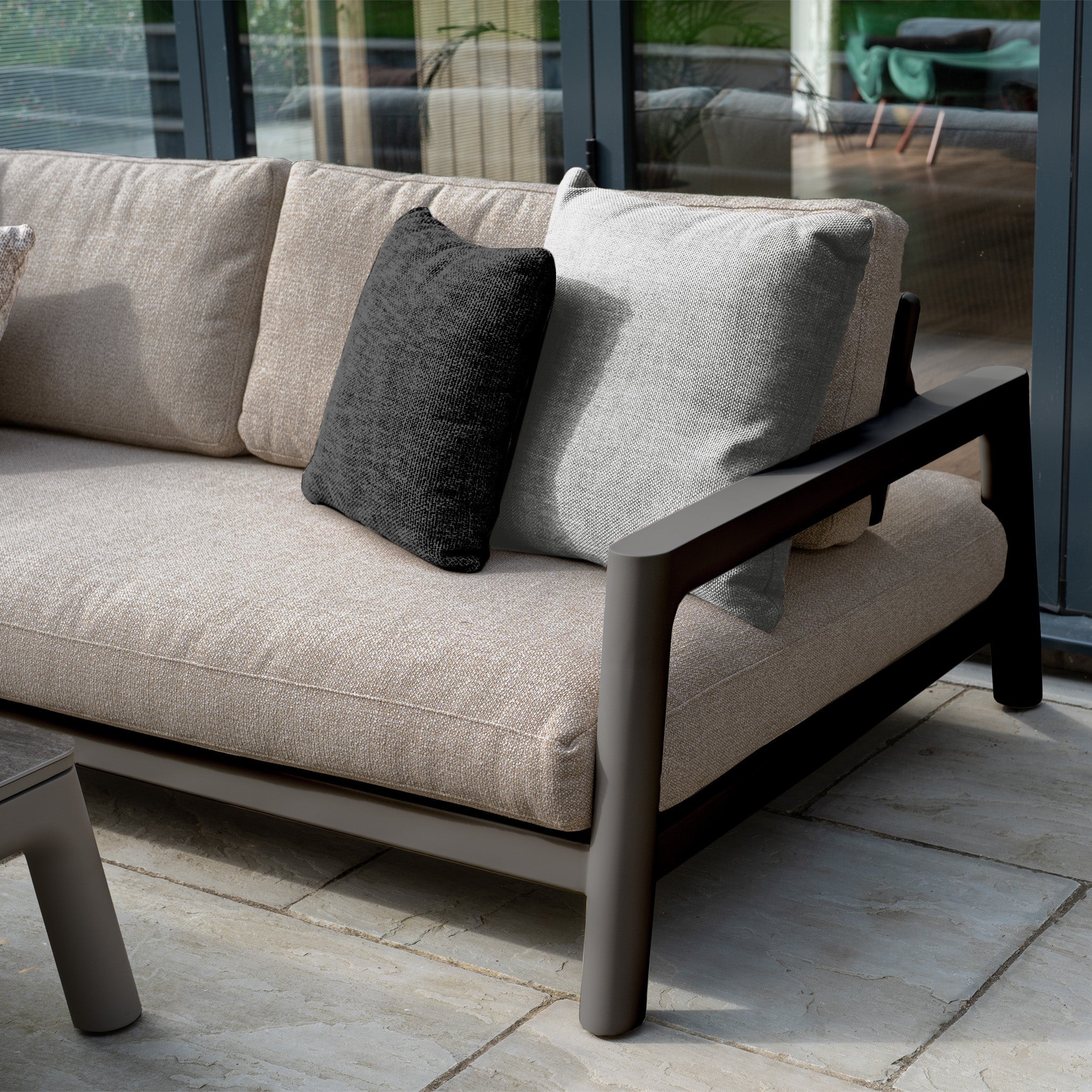 The Theia 3 Seat Sofa Set in Truffle features beige cushions and black and gray throw pillows, creating a modern look on any tiled patio near glass windows—perfect for stylish outdoor furniture arrangements.