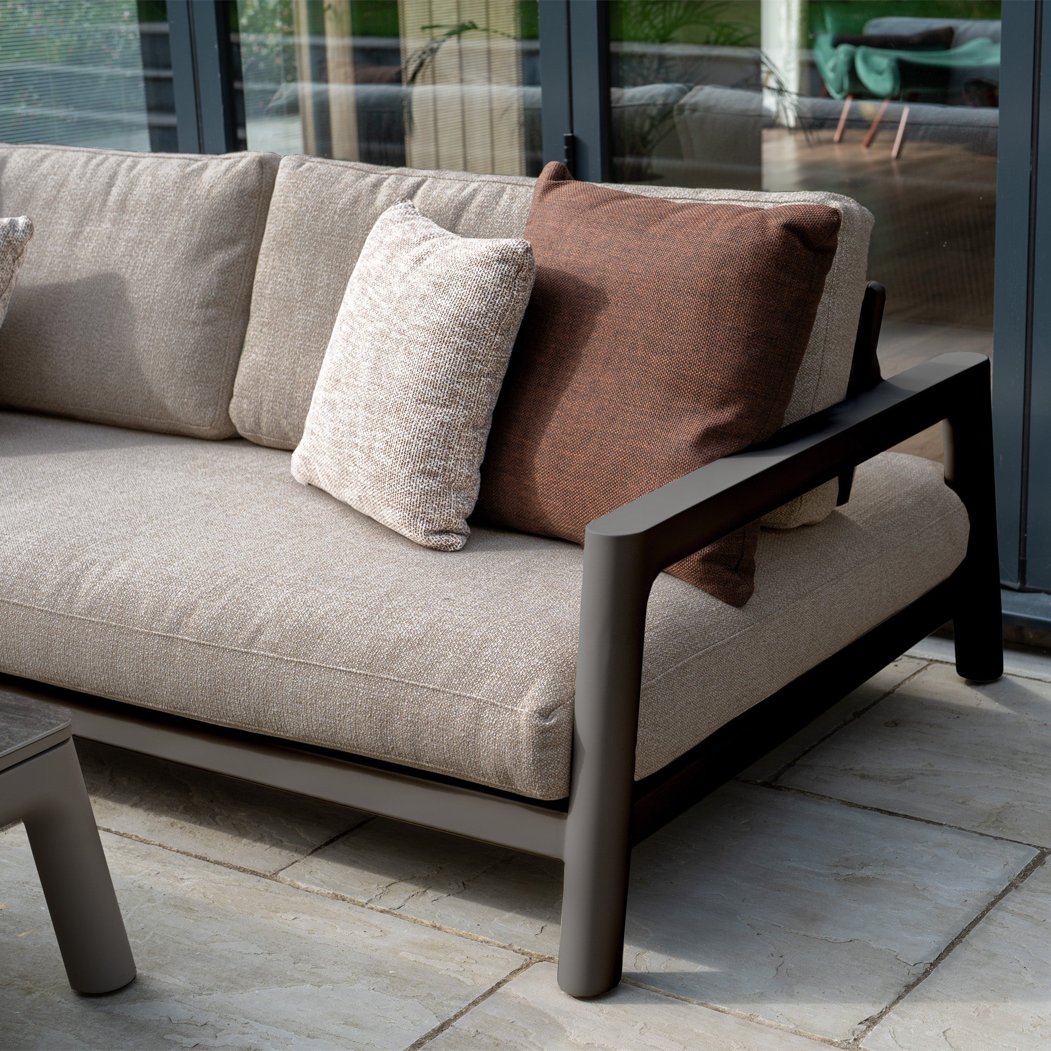 Theia 3 Seat Sofa Set in Truffle with brown and light beige cushions sits on a stone patio by a glass window—perfect outdoor furniture for stylish gatherings.