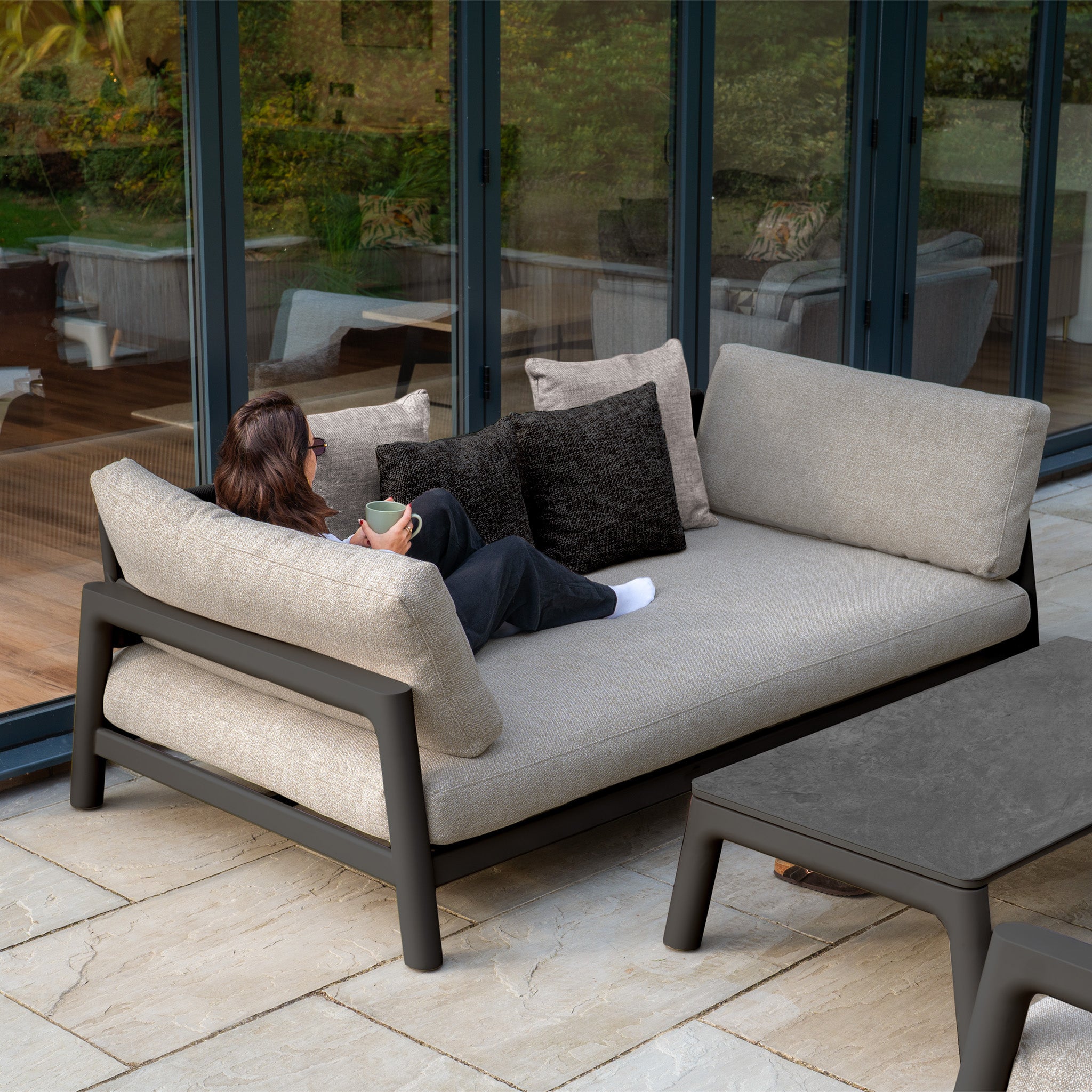 A person relaxes on the Theia 3 Seat Sofa Set in Truffle with pillows, holding a mug near glass doors and a table.