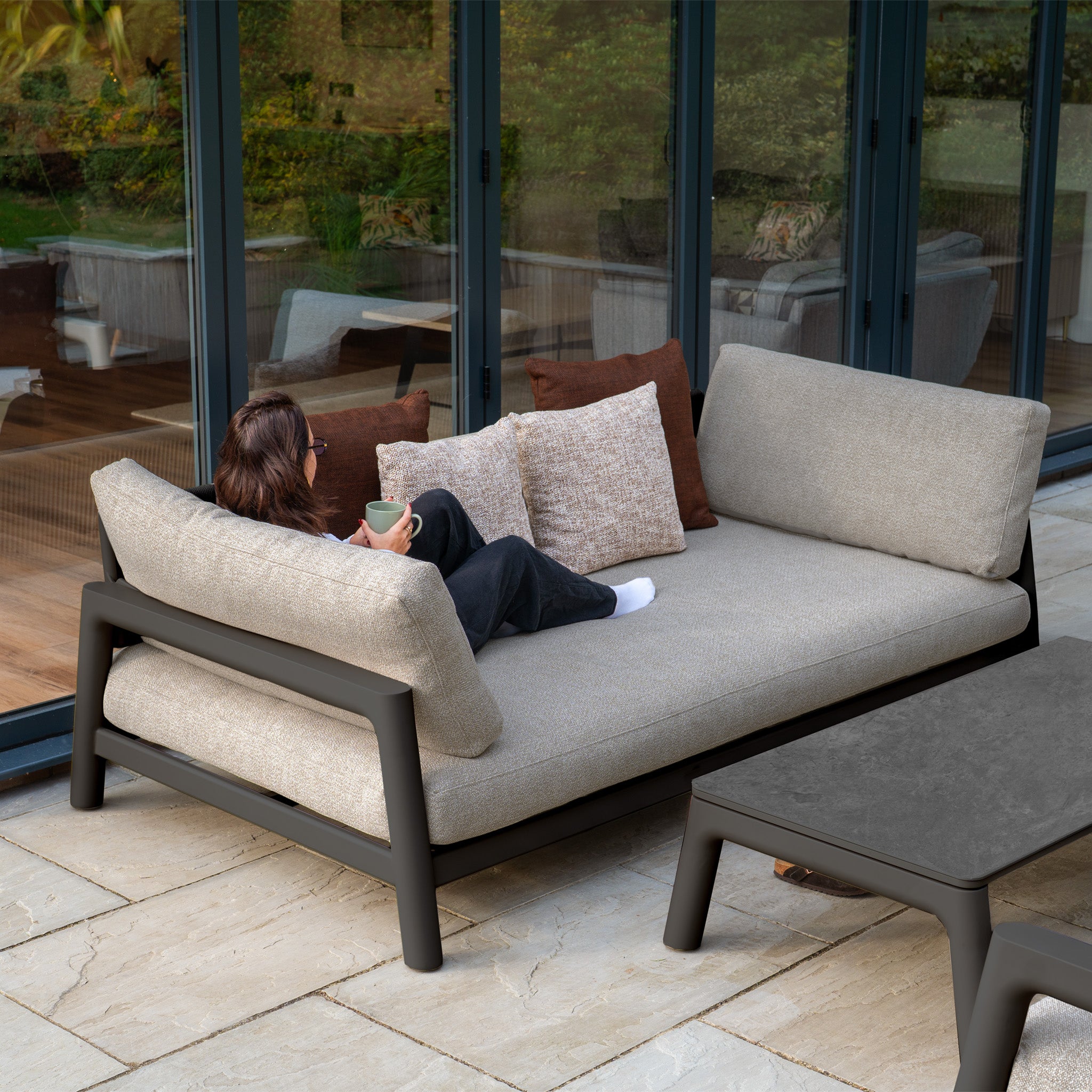 A woman relaxes with a mug on the Theia 3 Seat Sofa Set in Truffle, positioned outdoors facing large glass doors and a garden.