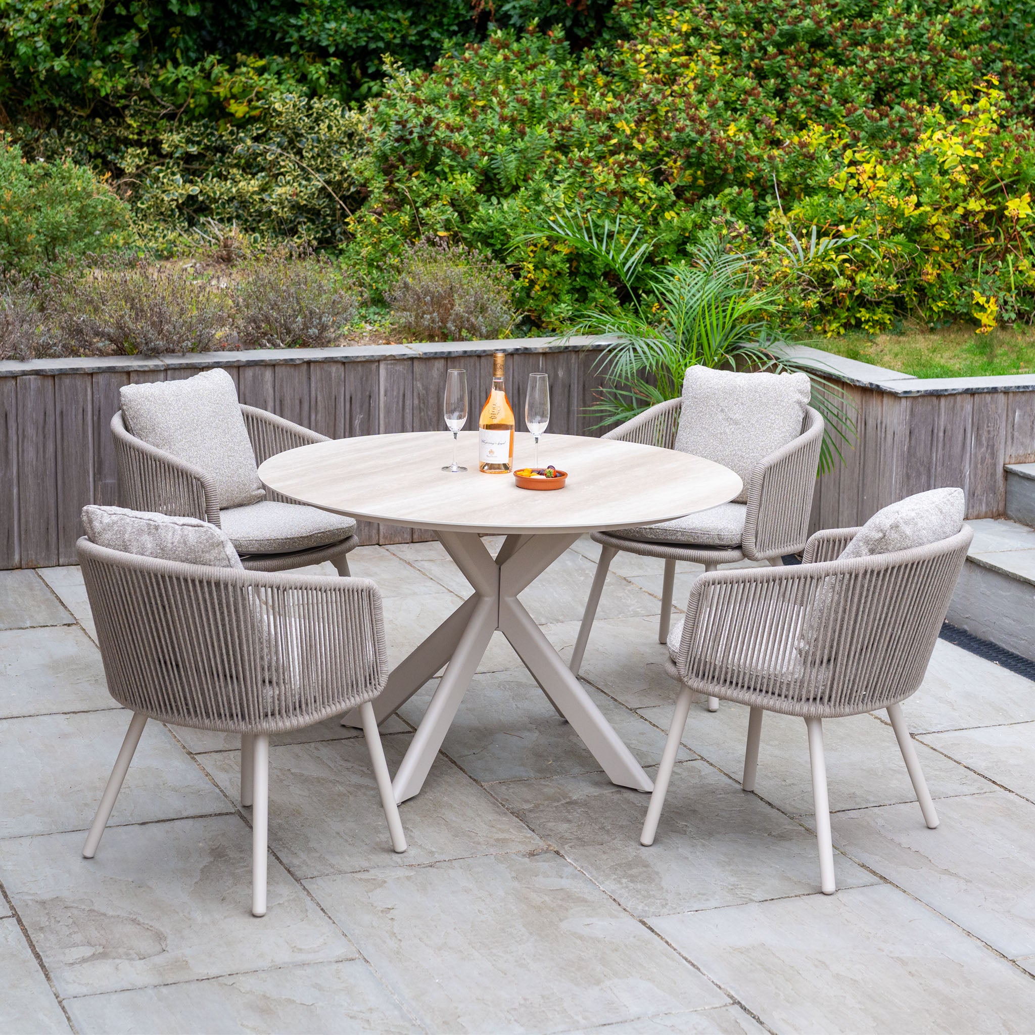 Enjoy outdoor dining with the Theia 4 Seat Round Rope Dining Set with Albatross Table in Oatmeal—four cushioned chairs and a stylish patio table, perfect for alfresco meals surrounded by lush greenery.