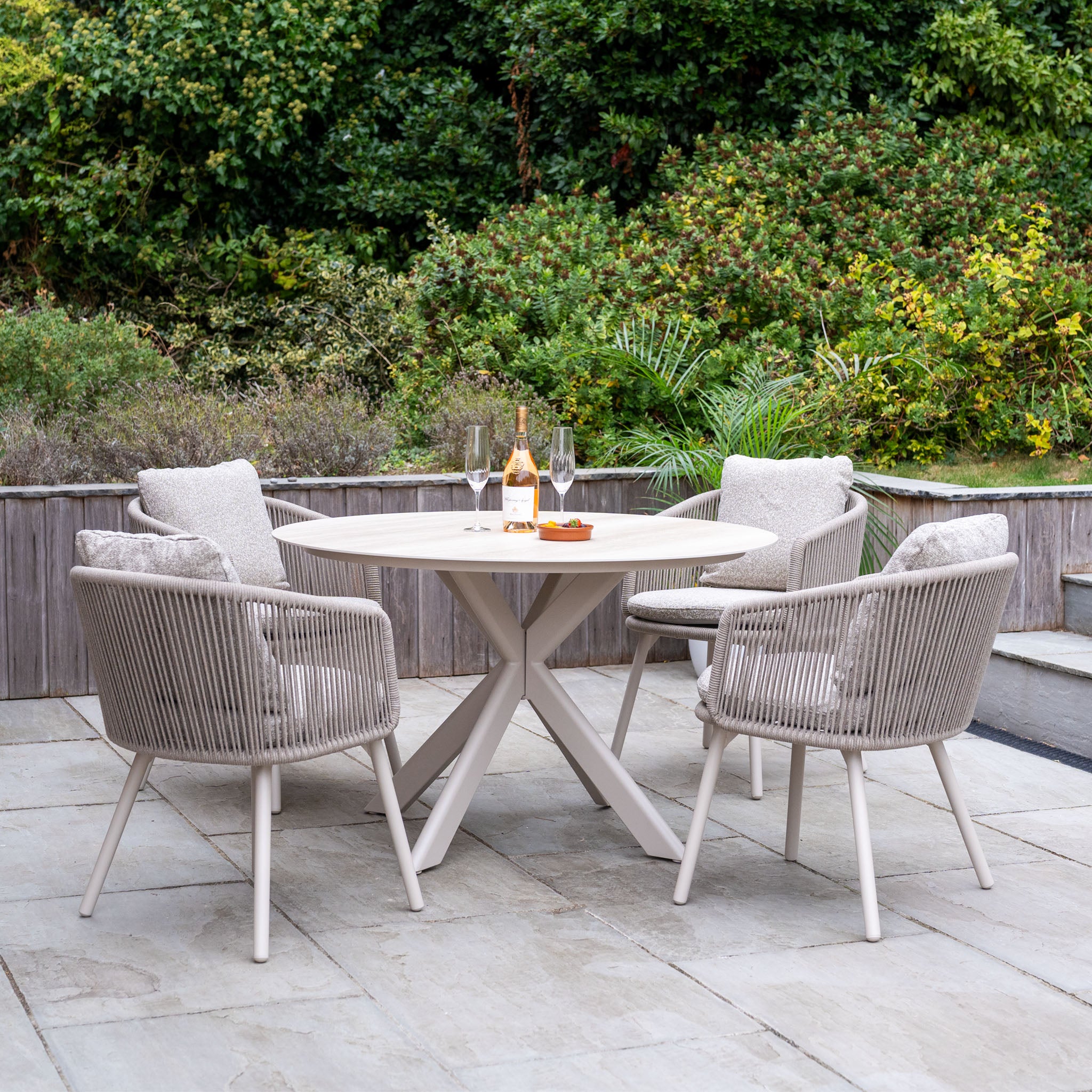 The Theia 4 Seat Round Rope Dining Set with Albatross Table in Oatmeal features cushioned chairs on a stone terrace, surrounded by greenery—ideal for relaxed outdoor dining.