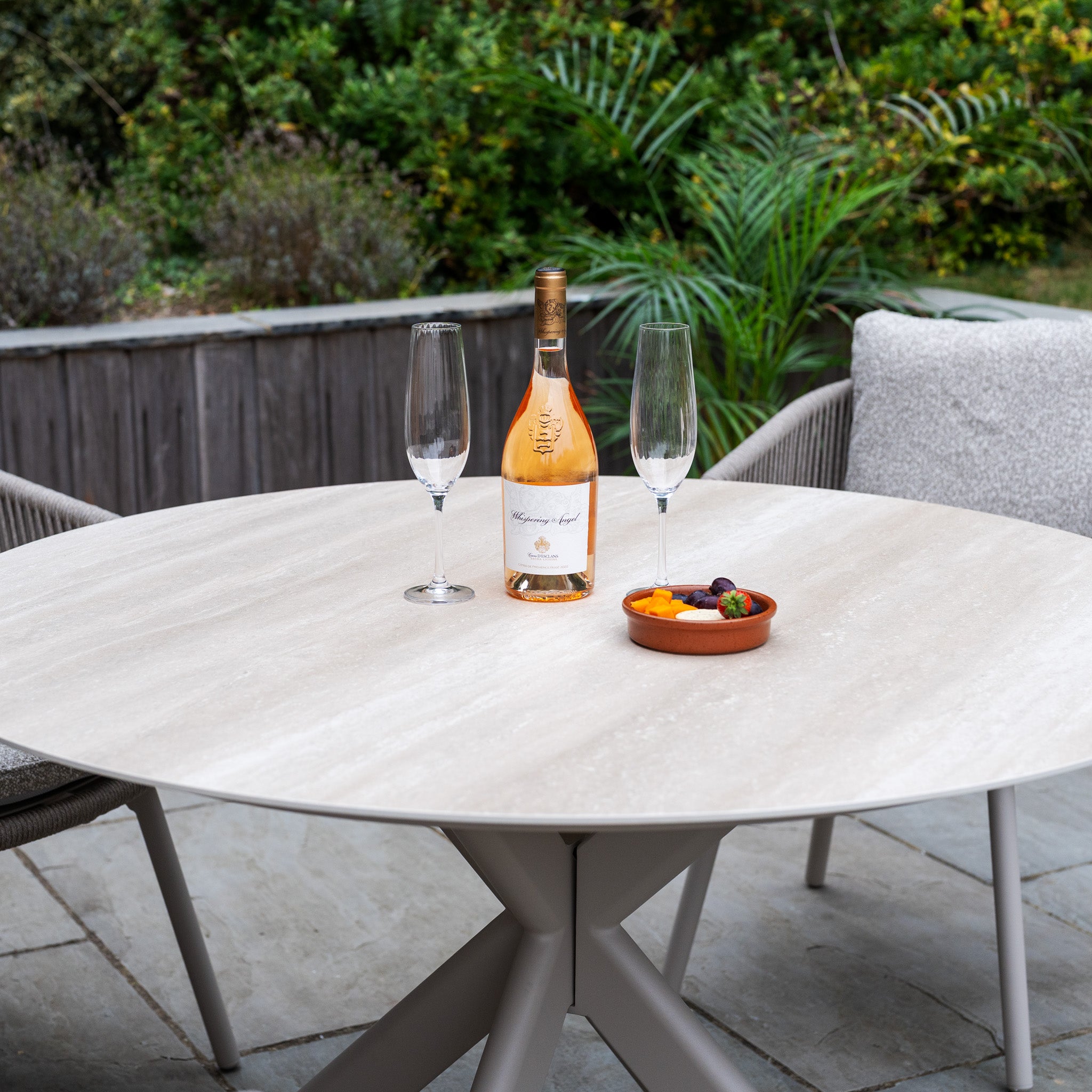 The Theia 4 Seat Round Rope Dining Set with Albatross Table in Oatmeal is set outside with rosé wine, two champagne flutes, and a bowl of fruit—ideal for stylish gatherings with Oatmeal Garden Furniture.
