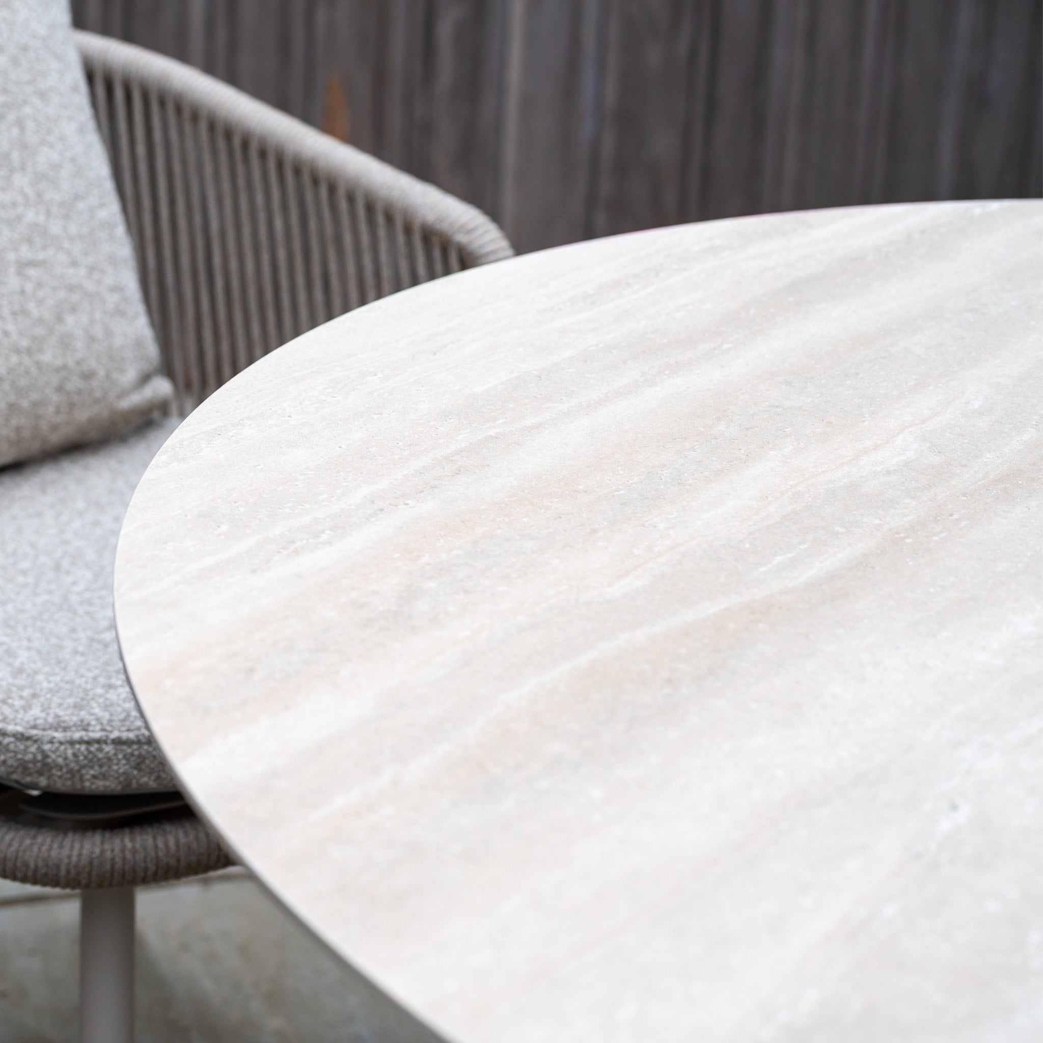 The Theia 4 Seat Round Rope Dining Set with Albatross Table in Oatmeal features a round stone table beside a cushioned chair, set elegantly outdoors with a wooden fence in the background.