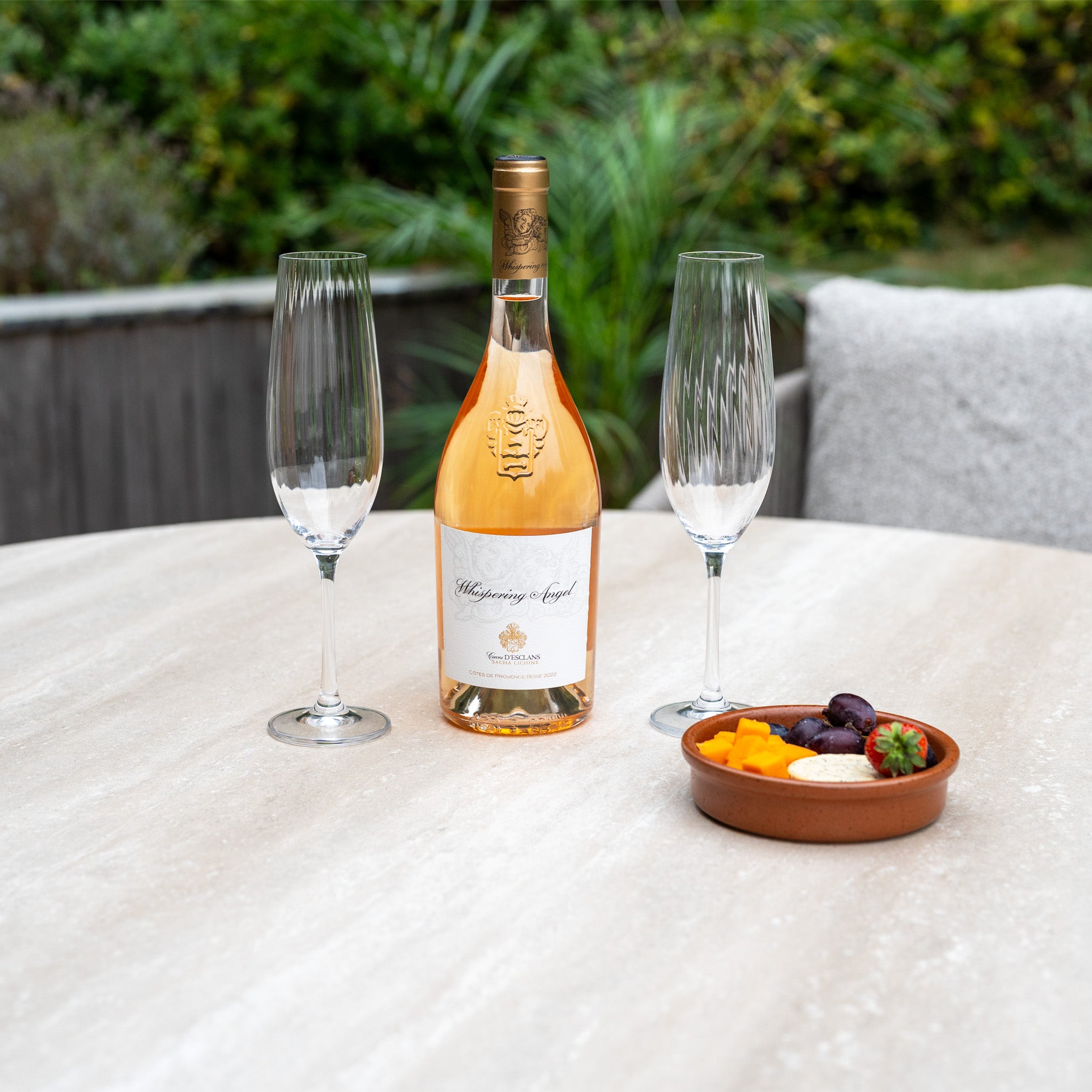 A bottle of rosé, two glasses, and snacks sit ready on an outdoor table—perfect for relaxing with the Theia 4 Seat Round Rope Dining Set with Albatross Table in Oatmeal.